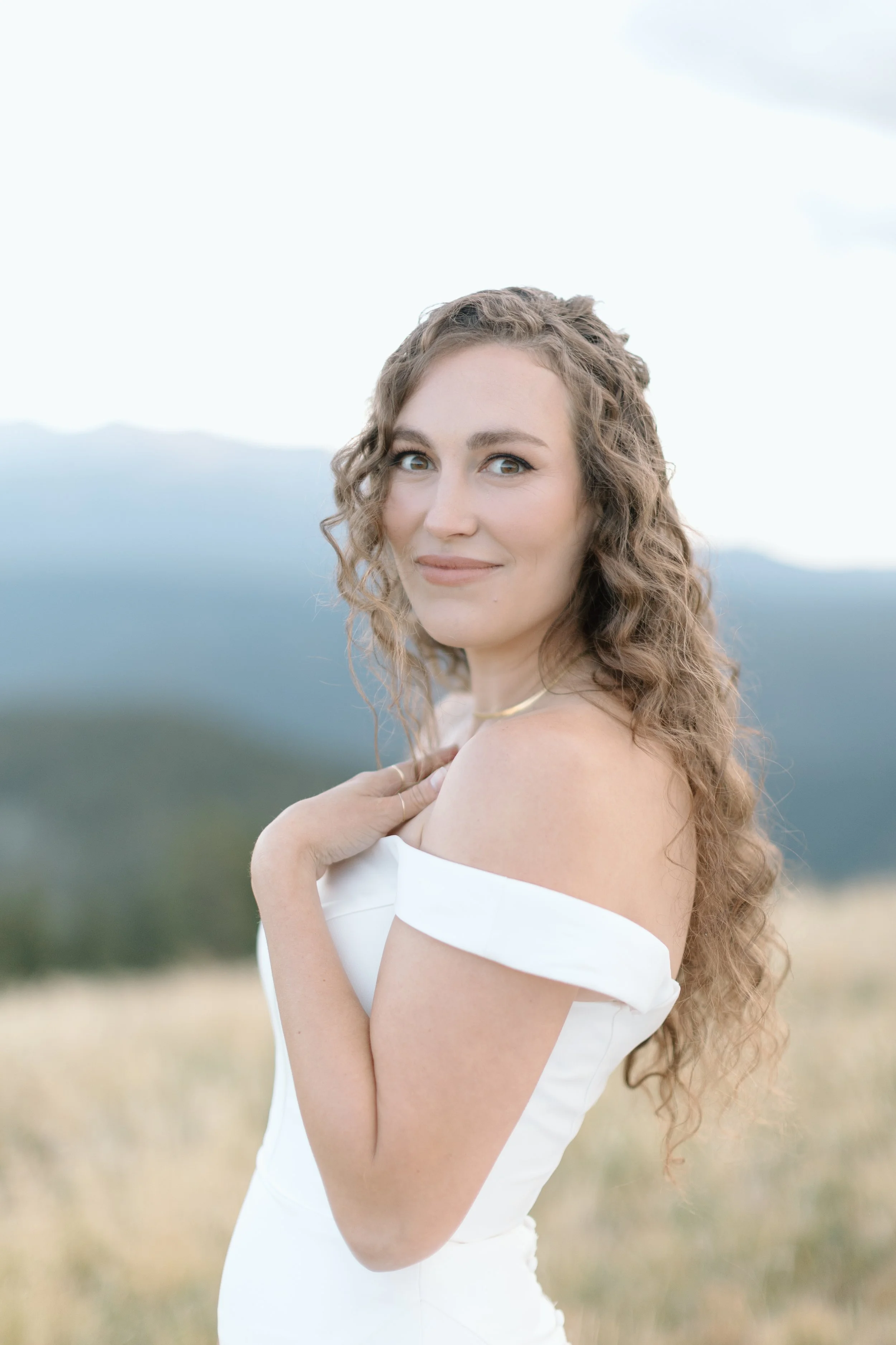 A woman with curly hair wearing an off-shoulder white dress standing outdoors with a mountain landscape in the background.