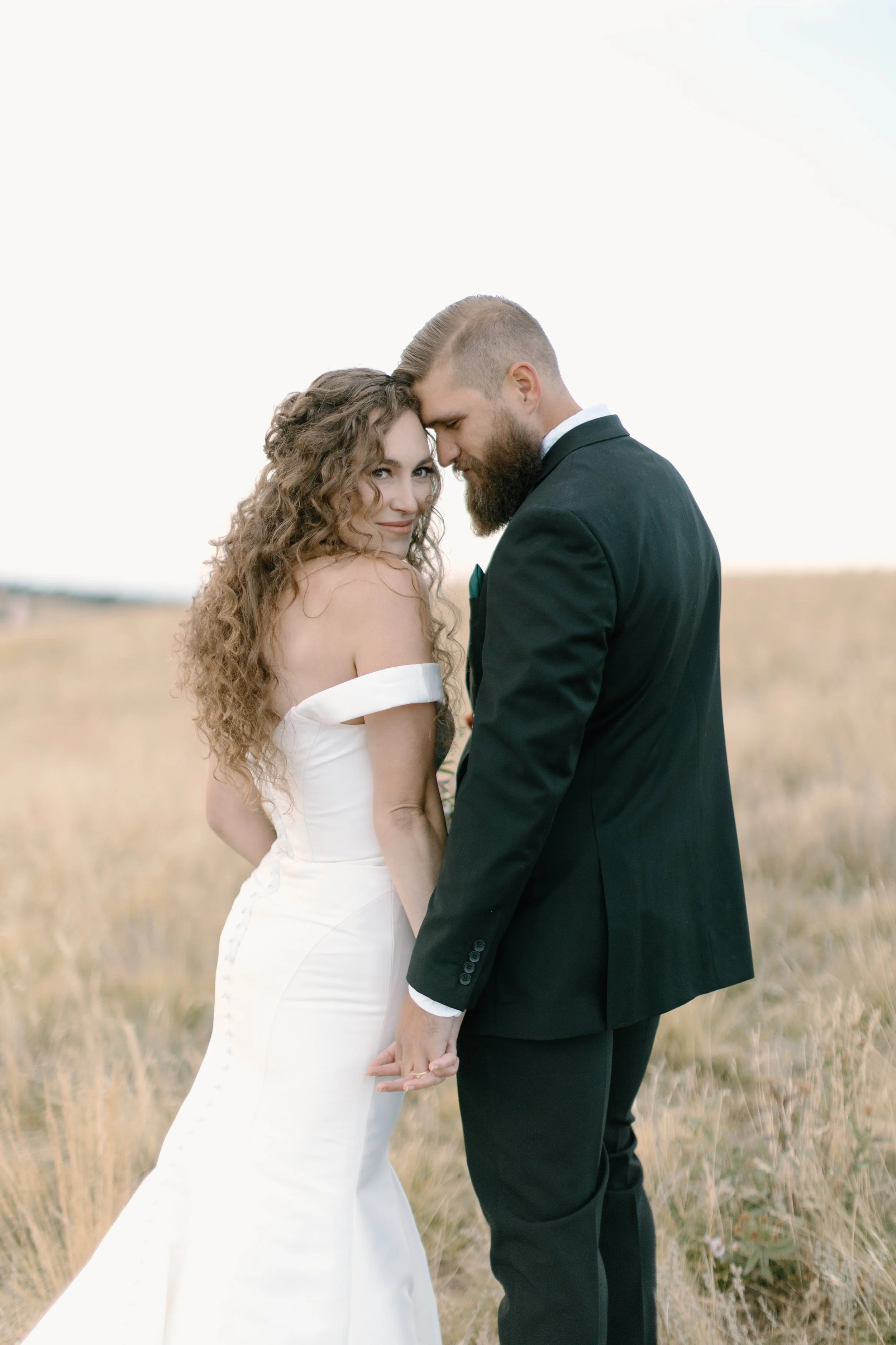 A bride and groom holding hands, standing in a field, with their foreheads touching and smiling at each other during their wedding shoot.