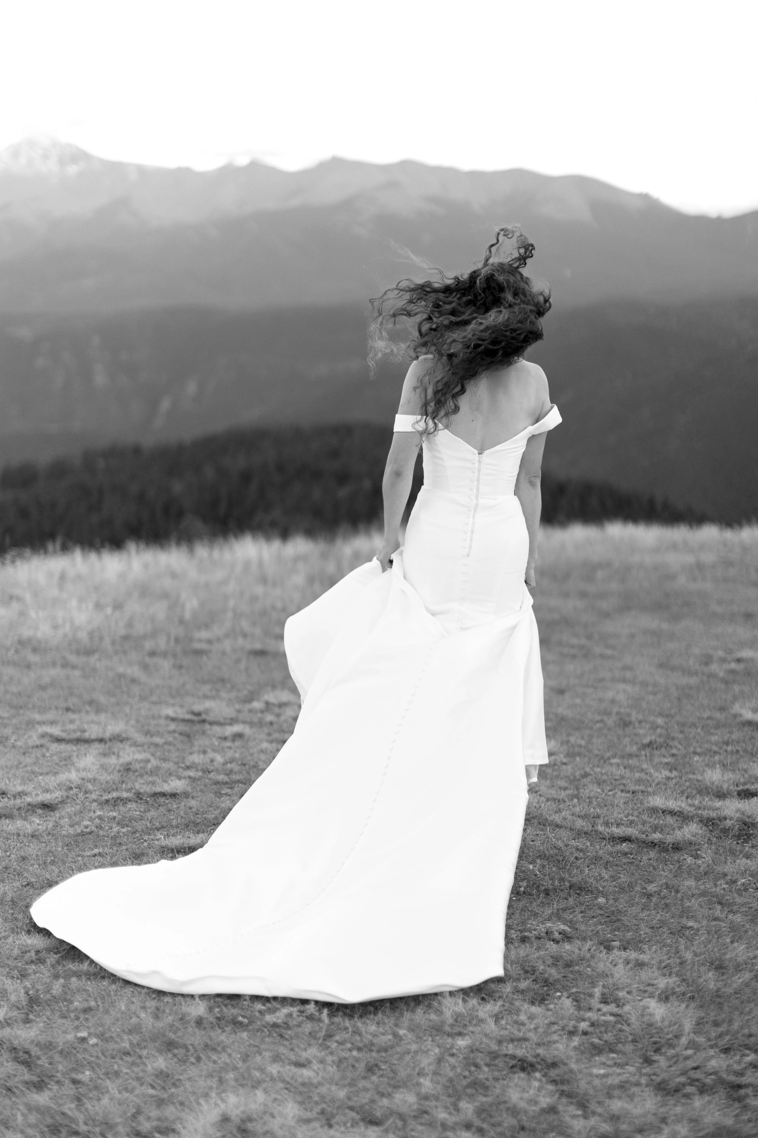 A woman in a wedding dress standing outdoors in a grassy field with mountains in the background, looking away from the camera.