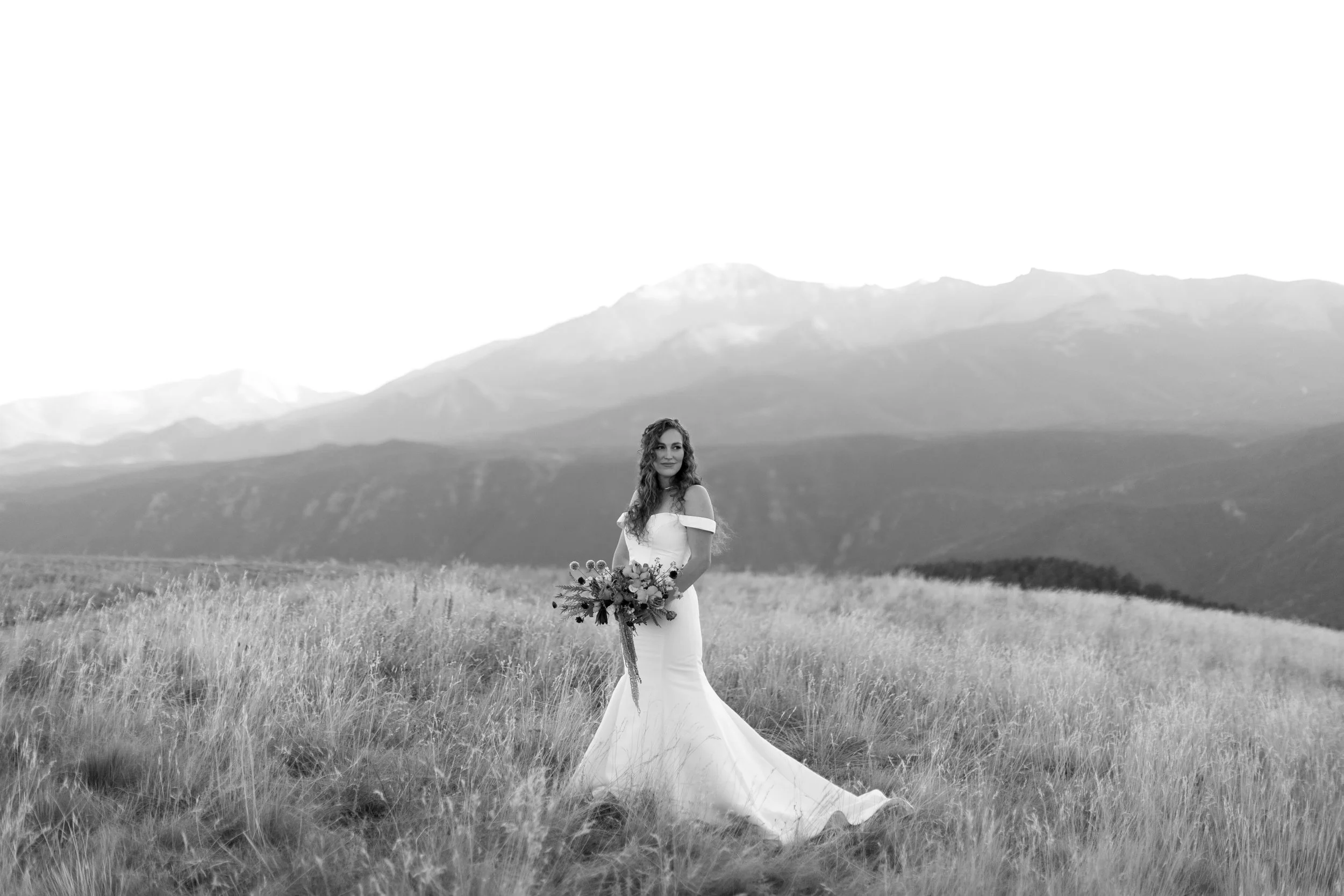 A bride in a wedding dress holding a bouquet, standing in a grassy field with mountains in the background.