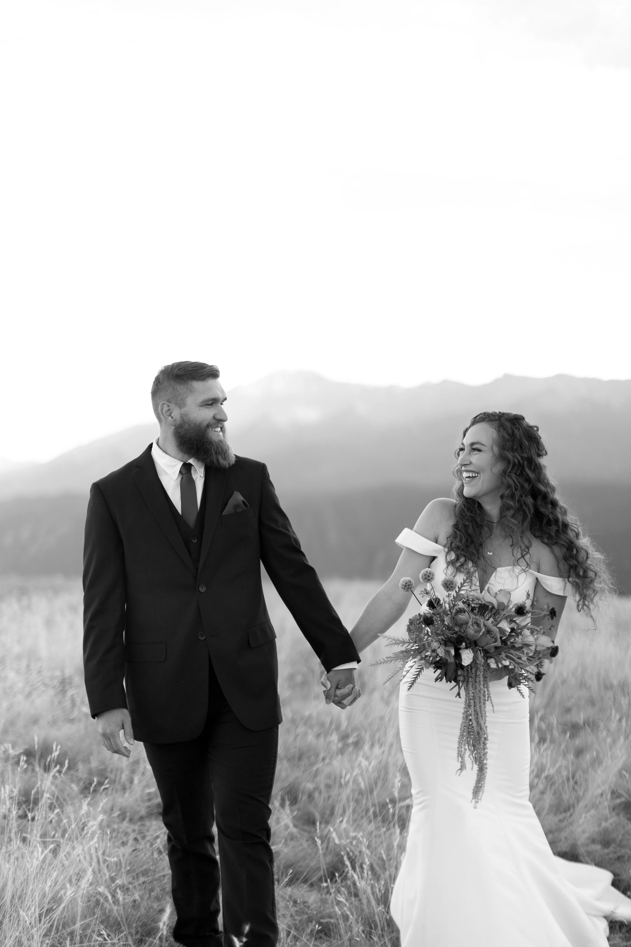 A black-and-white photo of a man and woman in wedding attire walking hand in hand through a grassy field with mountains in the background, both smiling and looking at each other.