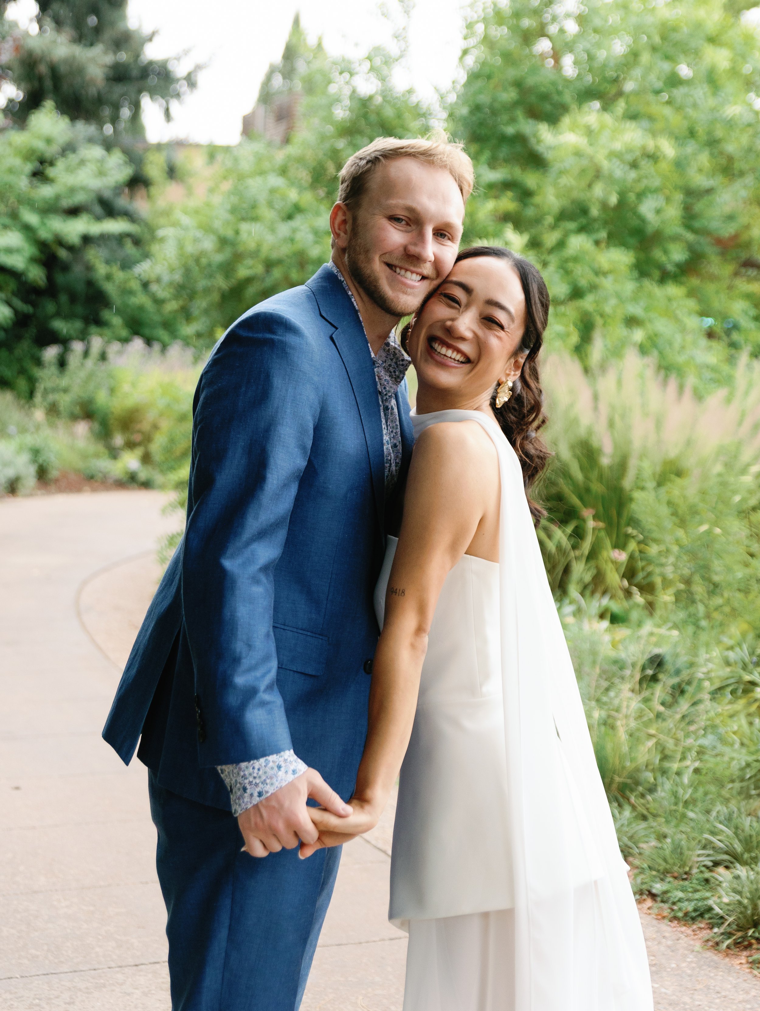 A happy couple, a man in a blue suit and a woman in a white dress, embrace and smile outdoors with greenery in the background.