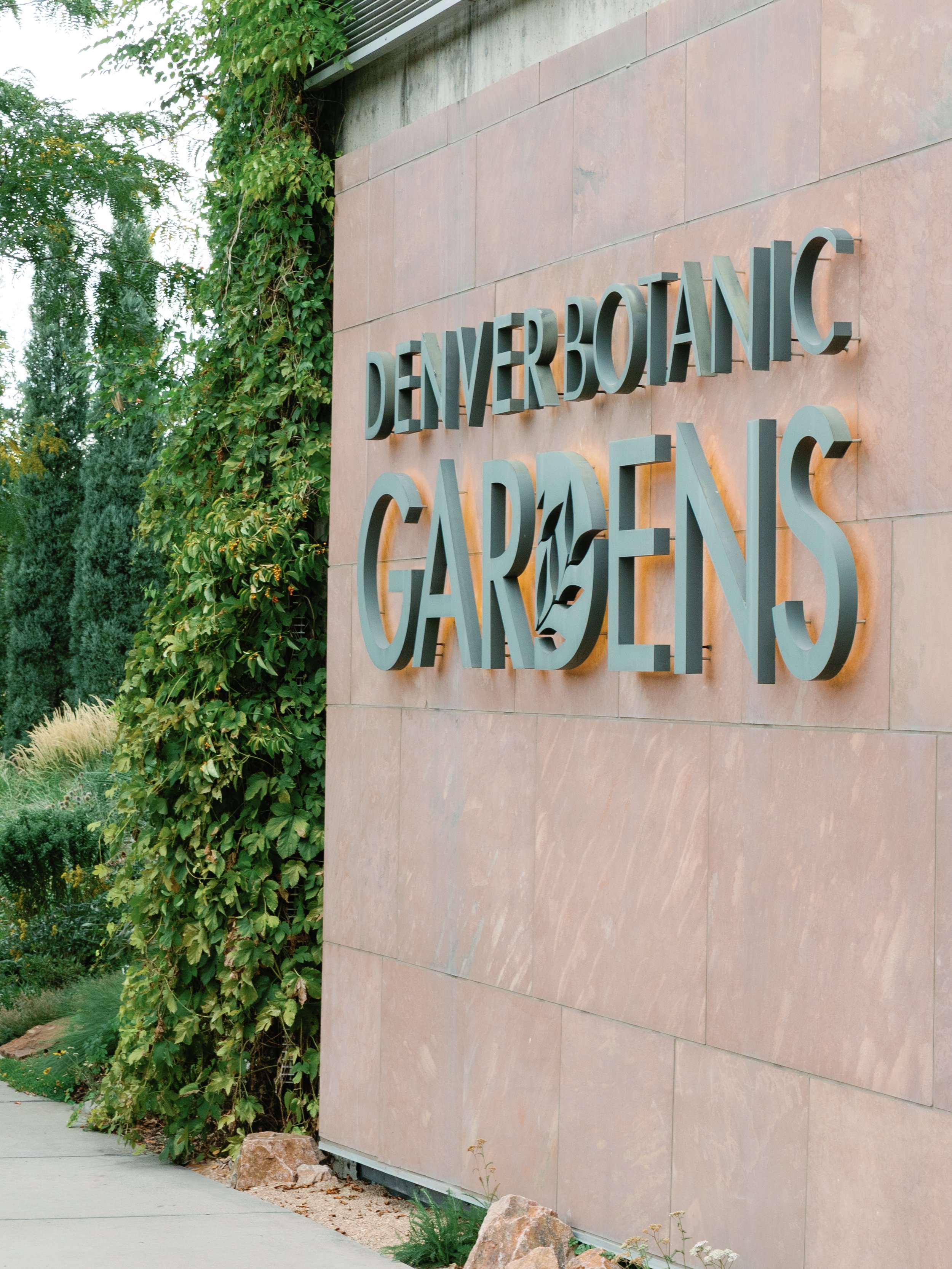Sign on a brick wall reading 'Denver Botanic Gardens' with greenery and trees around.