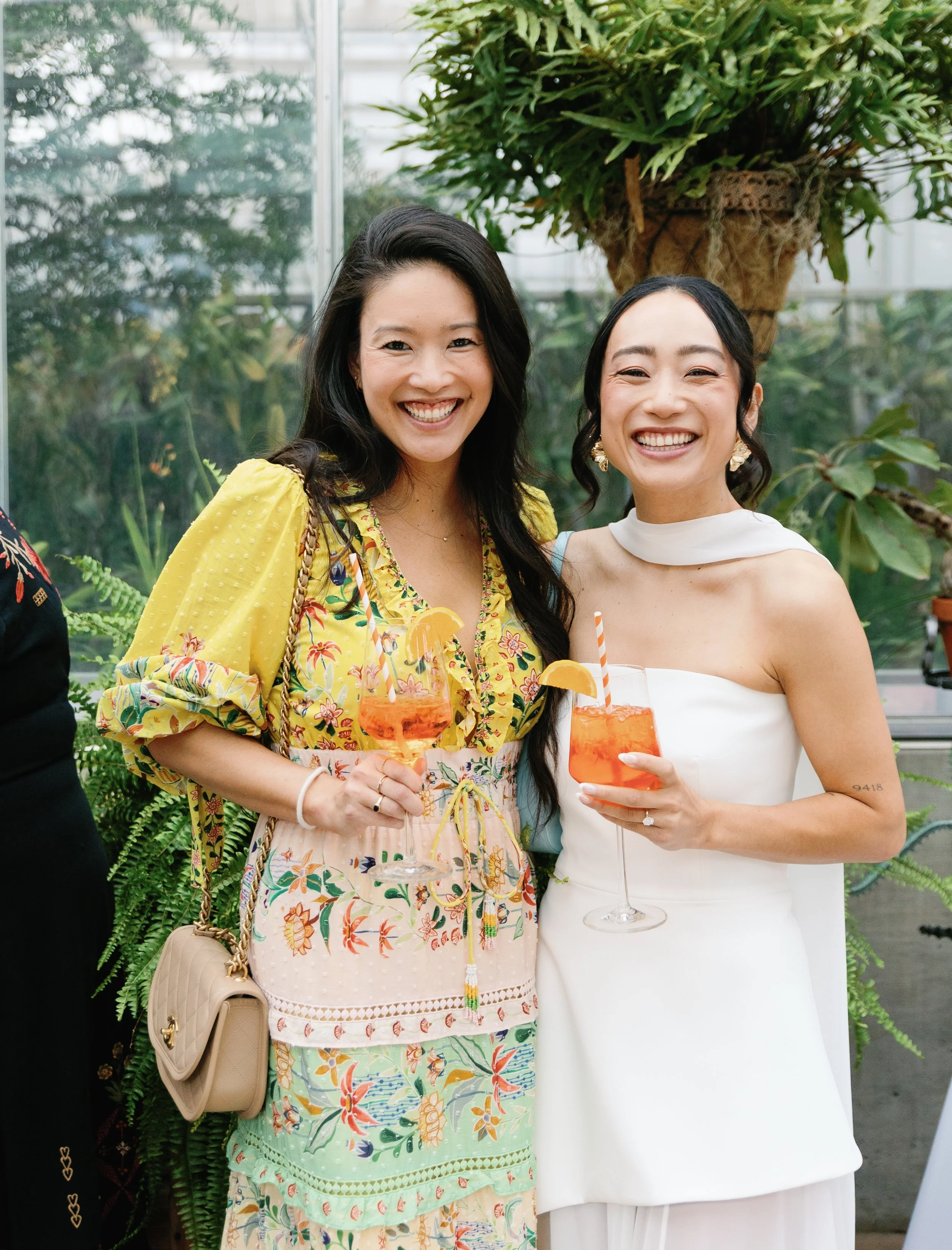 Two smiling women holding cocktail glasses with orange slices, posing indoors with lush green plants in the background.