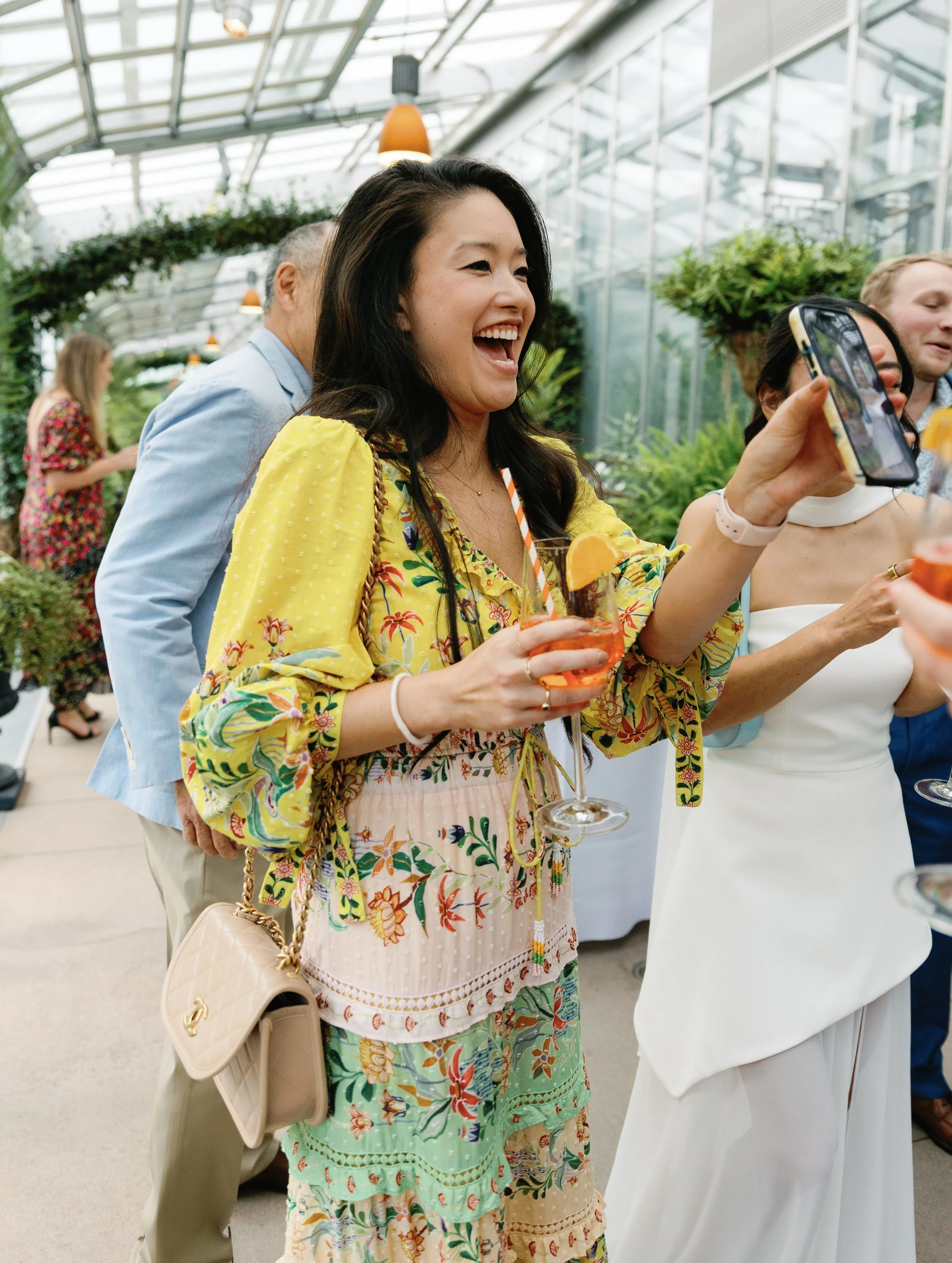 A woman wearing a colorful, floral dress is laughing and taking a selfie with her smartphone at a social gathering inside a greenhouse or conservatory, holding a cocktail with an orange slice garnish.