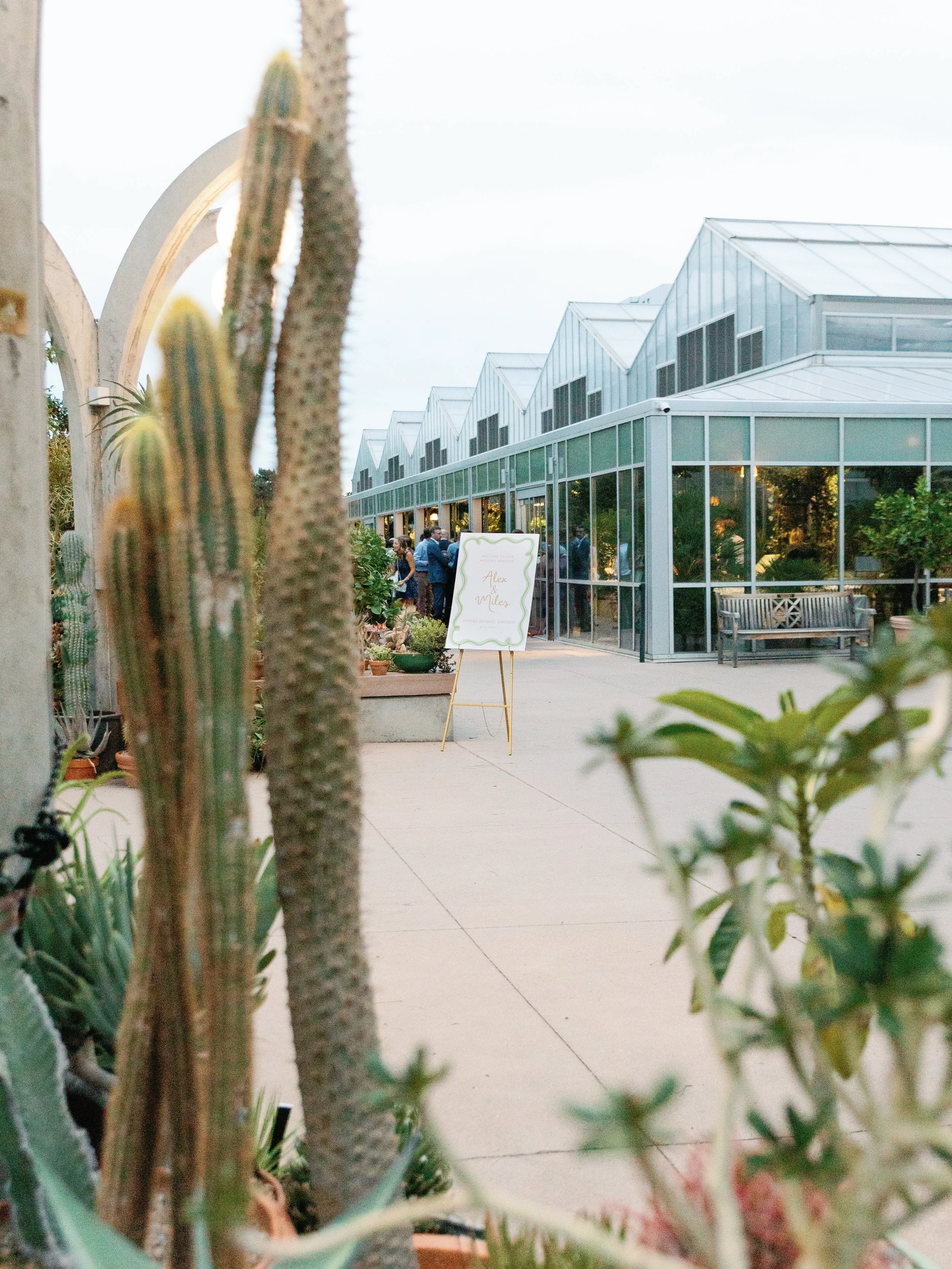 Greenhouse with glass walls and peaked roof, with a sign on a stand reading 'Alex & Miles,' surrounded by potted plants and cacti on a patio. People are gathered inside and outside the greenhouse.