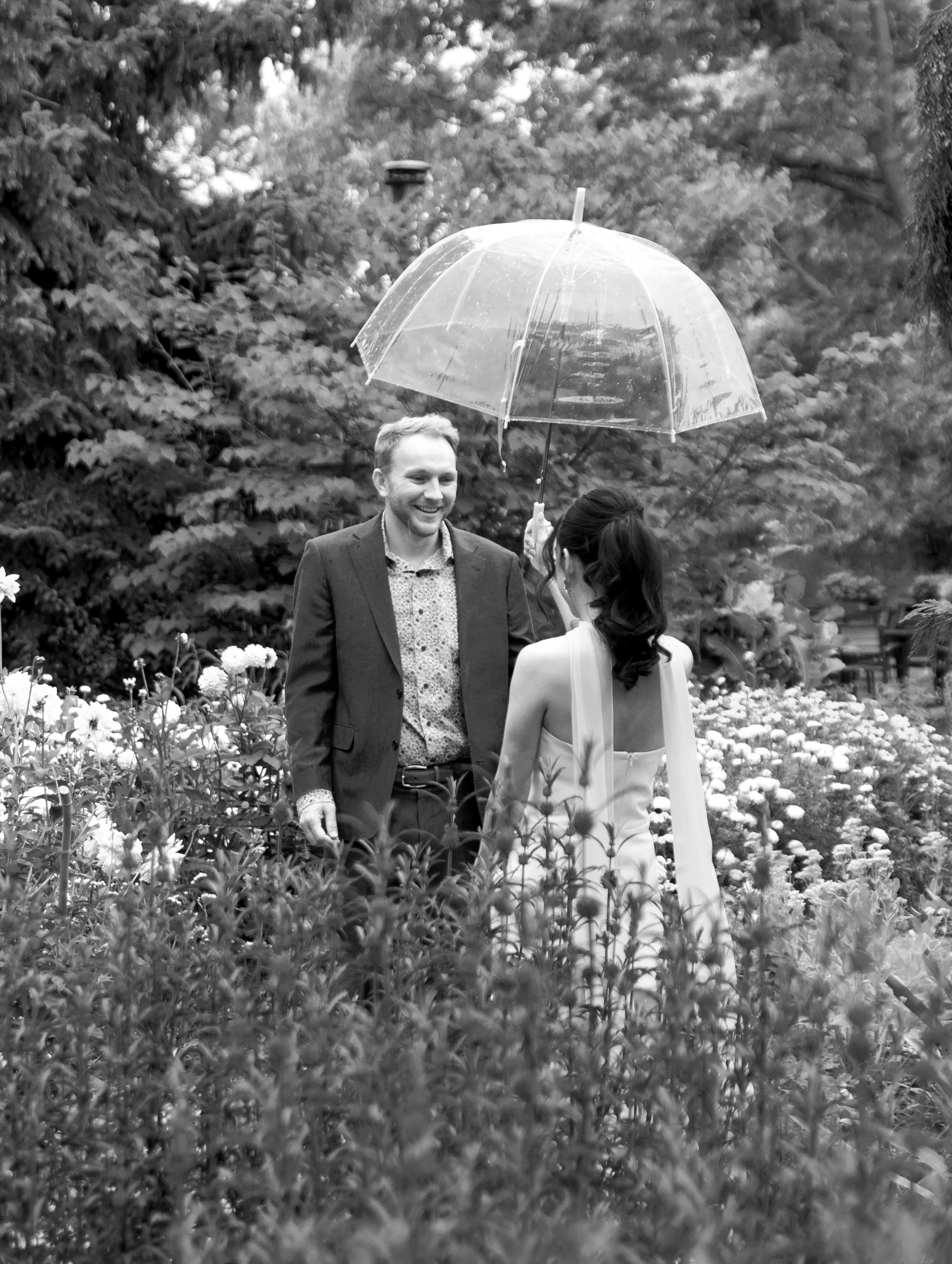 A man and woman standing in a garden, sharing a moment under a transparent umbrella, surrounded by flowers and trees.