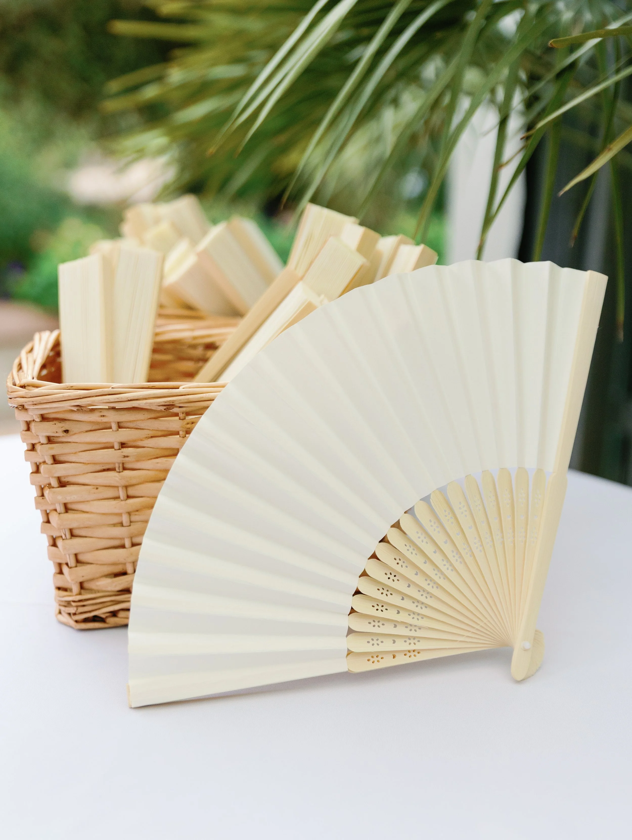 A white folding hand fan resting on a table with a wicker basket filled with wooden sticks in the background, outdoors with green foliage.