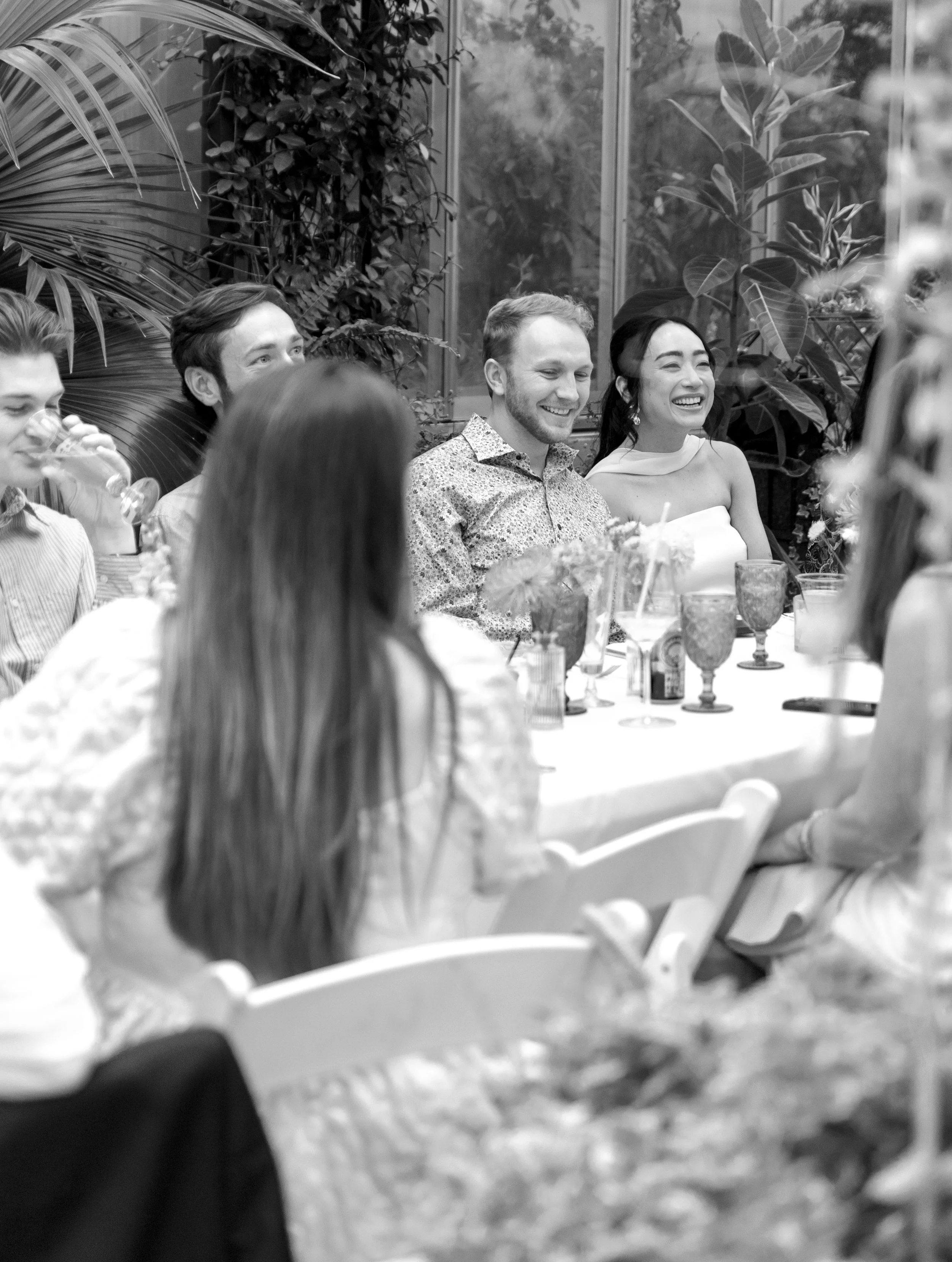 People sitting at a table enjoying a social gathering with drinks, smiling and laughing, in a well-lit indoor setting with large windows and lush plants in the background.