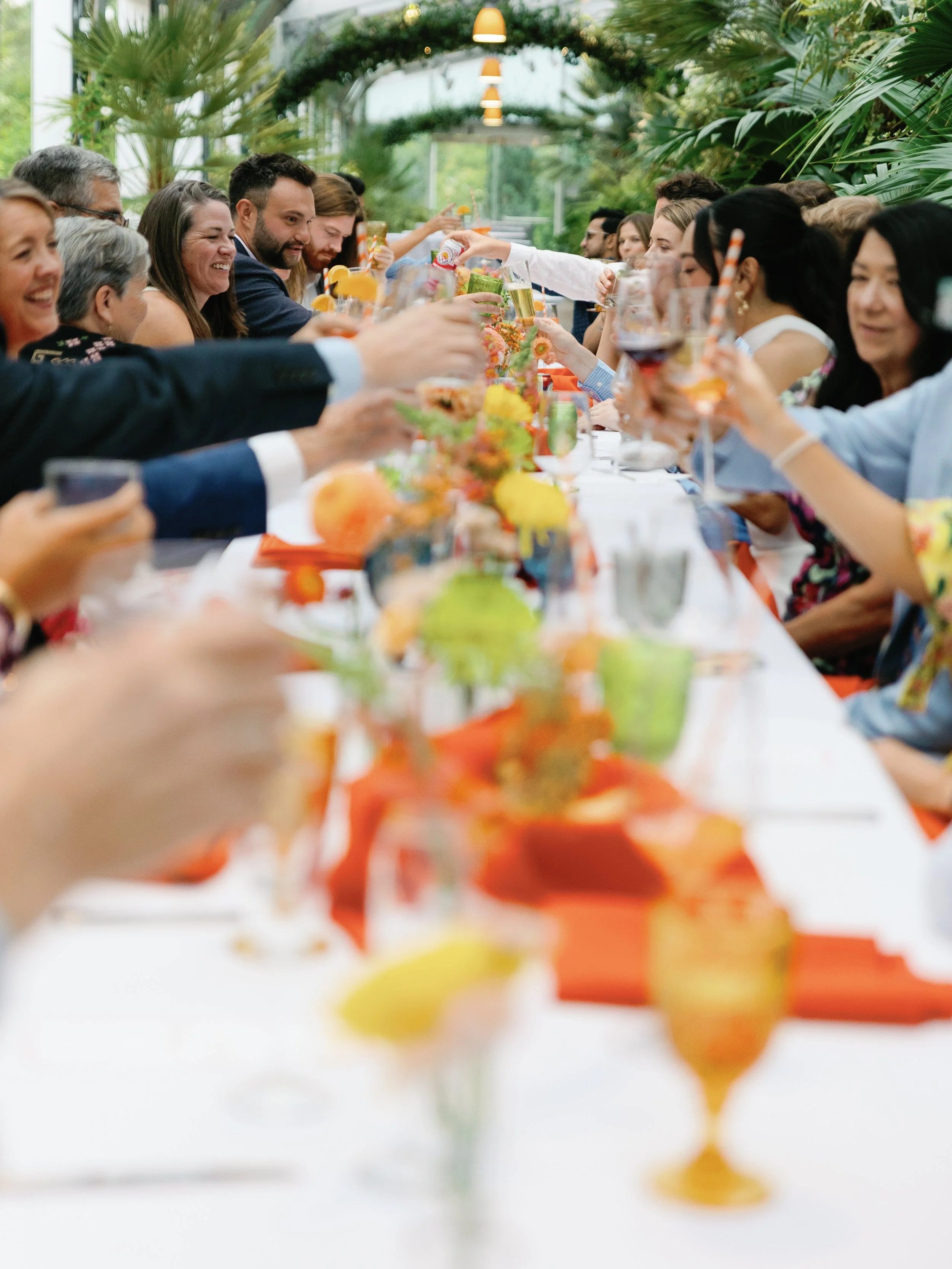 People at a long banquet table celebrating, toasting with drinks in a bright, indoor garden setting with plants and hanging lights.