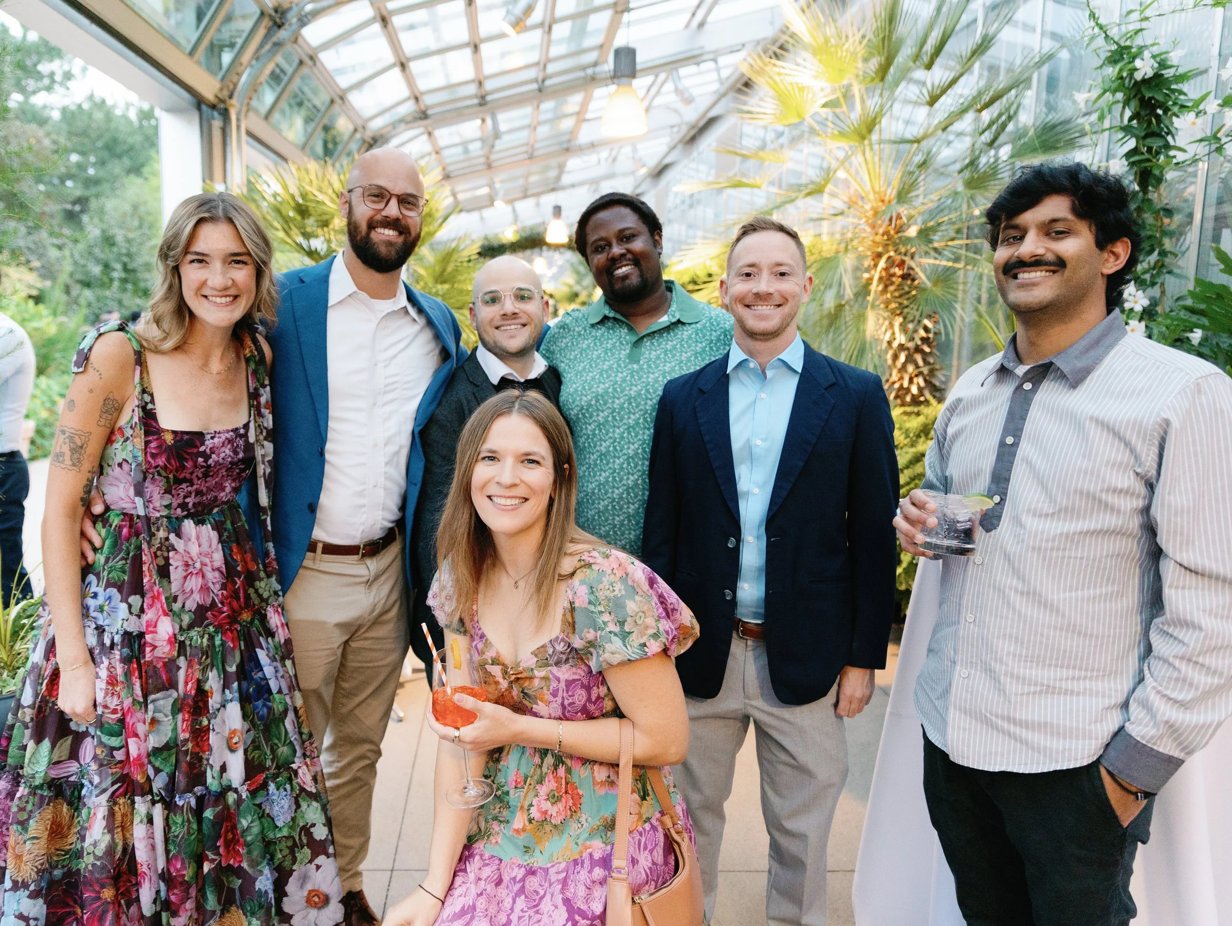 Group of seven diverse people smiling and posing together at a social event in a greenhouse with plants and glass ceiling.