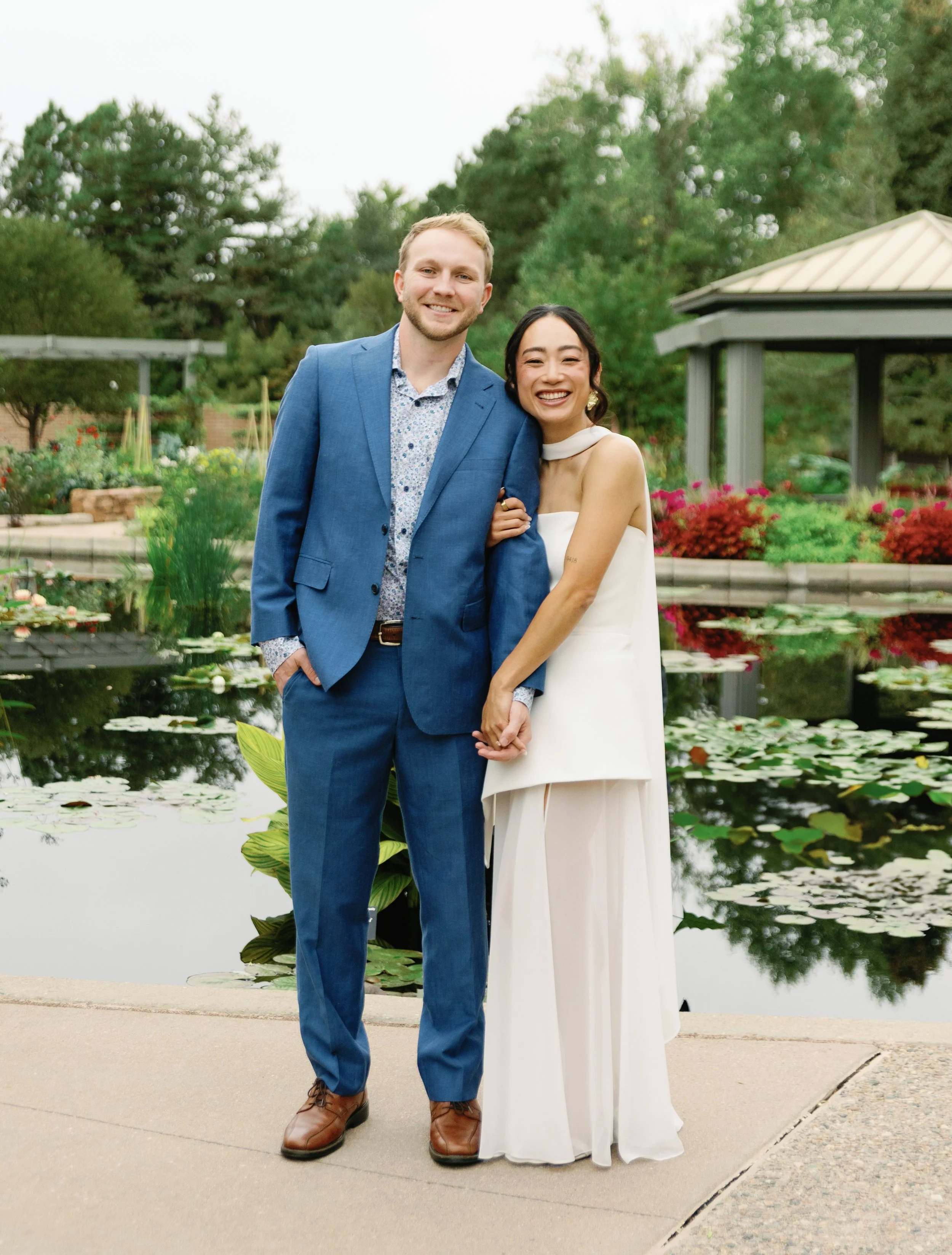 A smiling couple in formal attire, holding hands, standing by a pond with lily pads and colorful flowers, with greenery and a gazebo in the background.