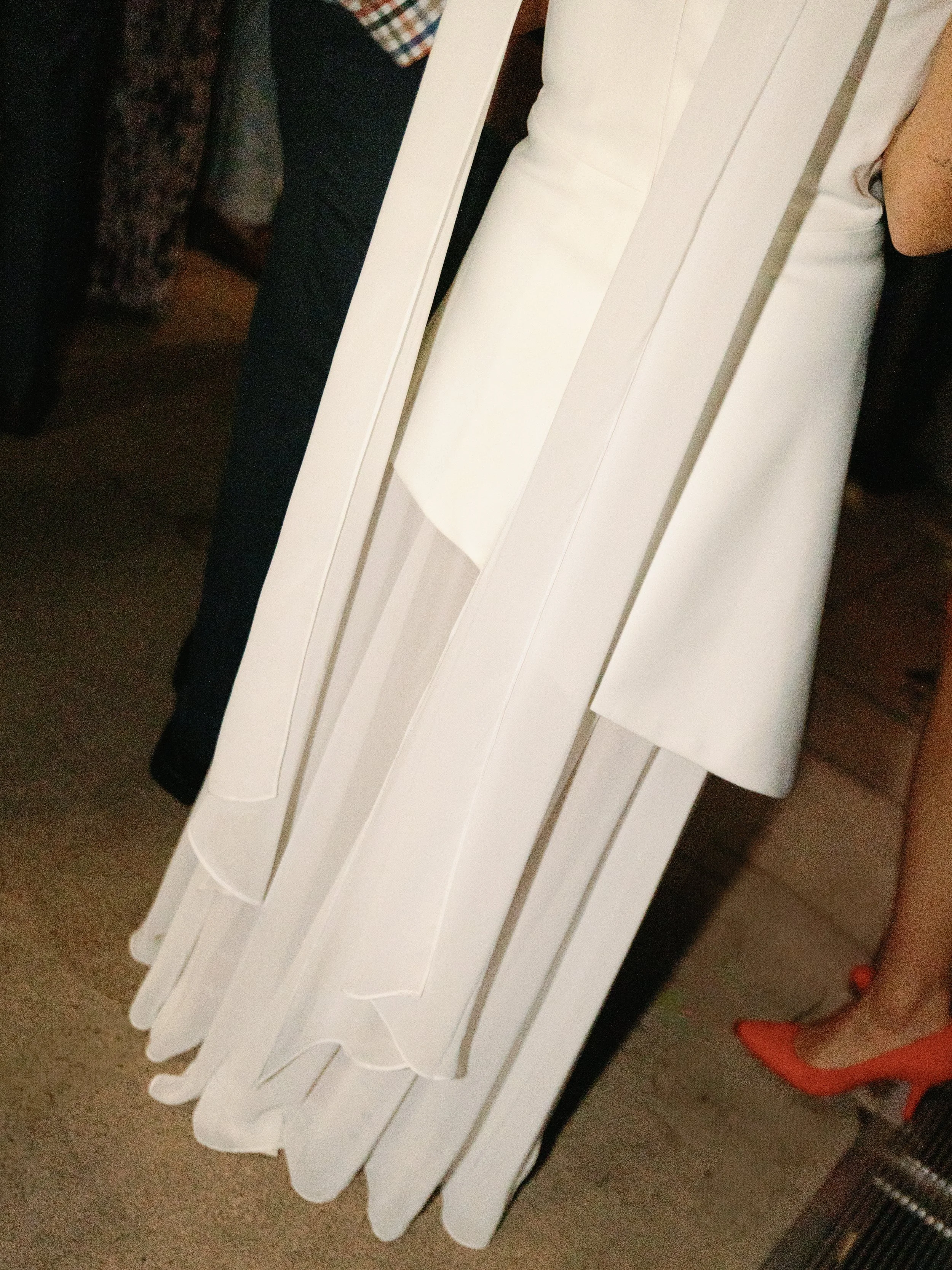 A person wearing a white dress or coat with pleated or layered fabric, standing on a wooden floor with other people nearby.