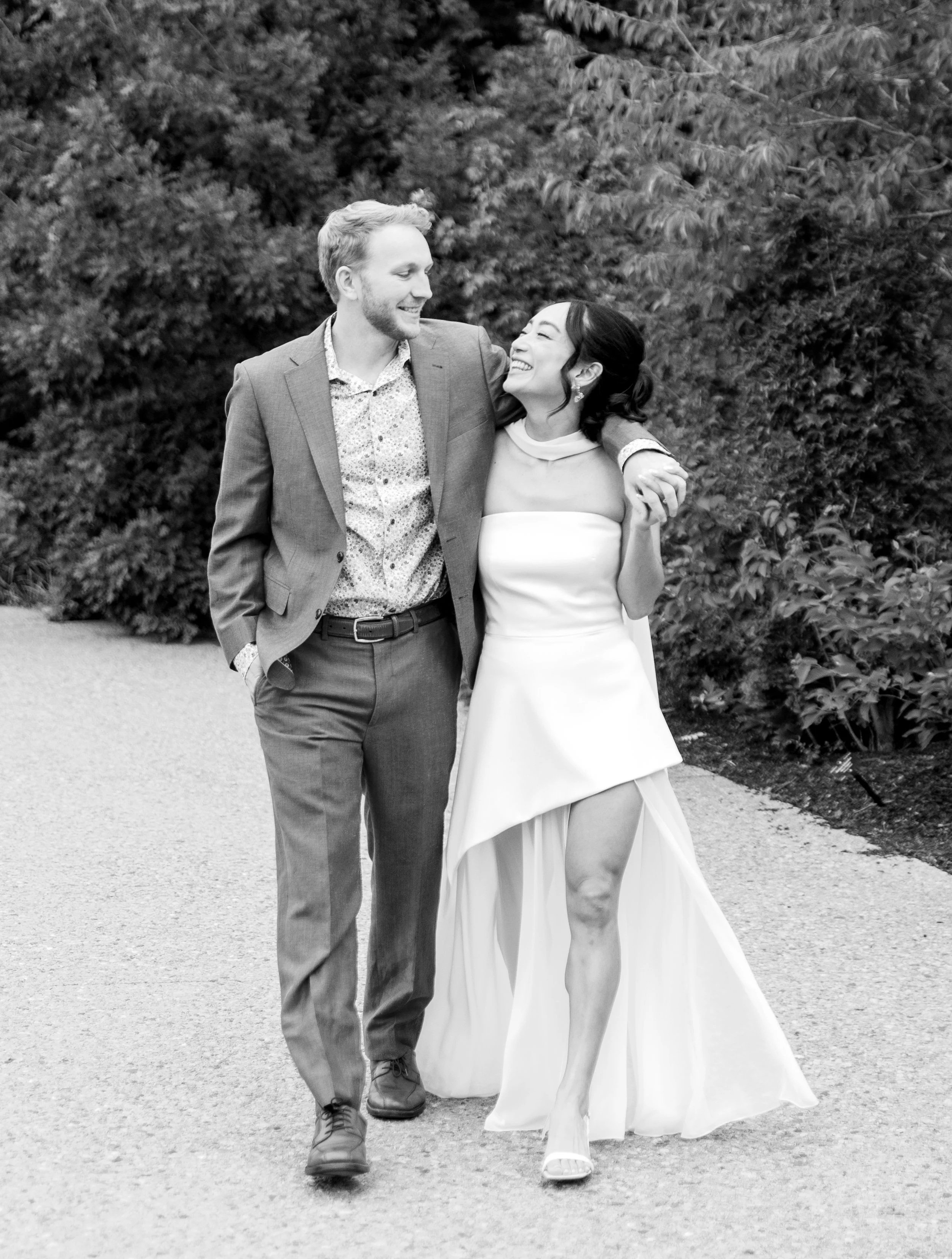 A happy couple, a man in a suit and a woman in a white dress, walking arm-in-arm outdoors with trees in the background.
