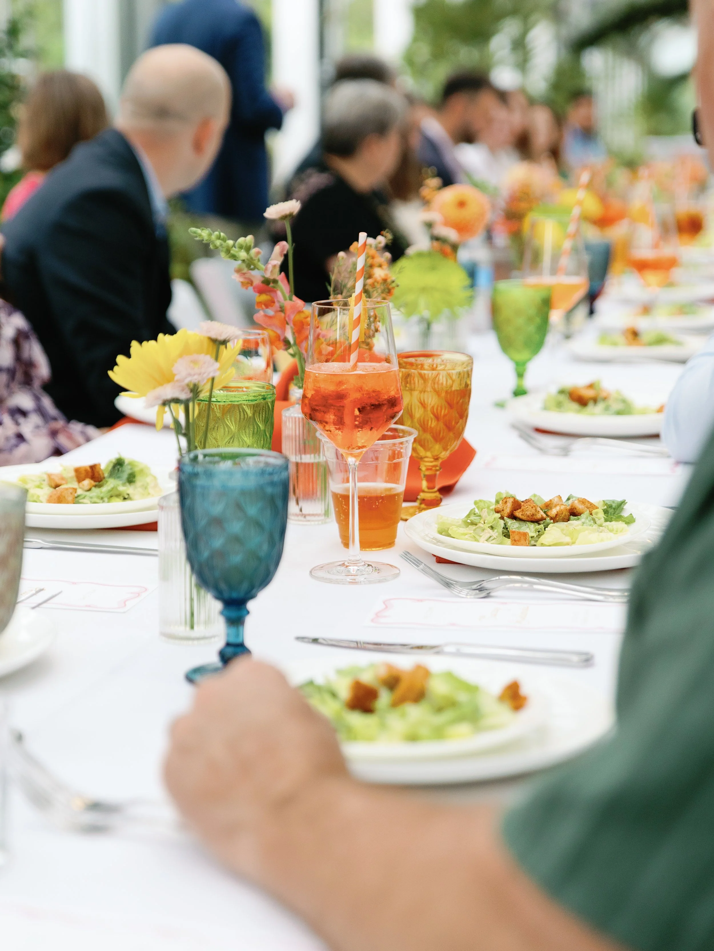 A table set for a social gathering, with colorful glasses, plates of salad, and floral decorations, outside at a daytime event.