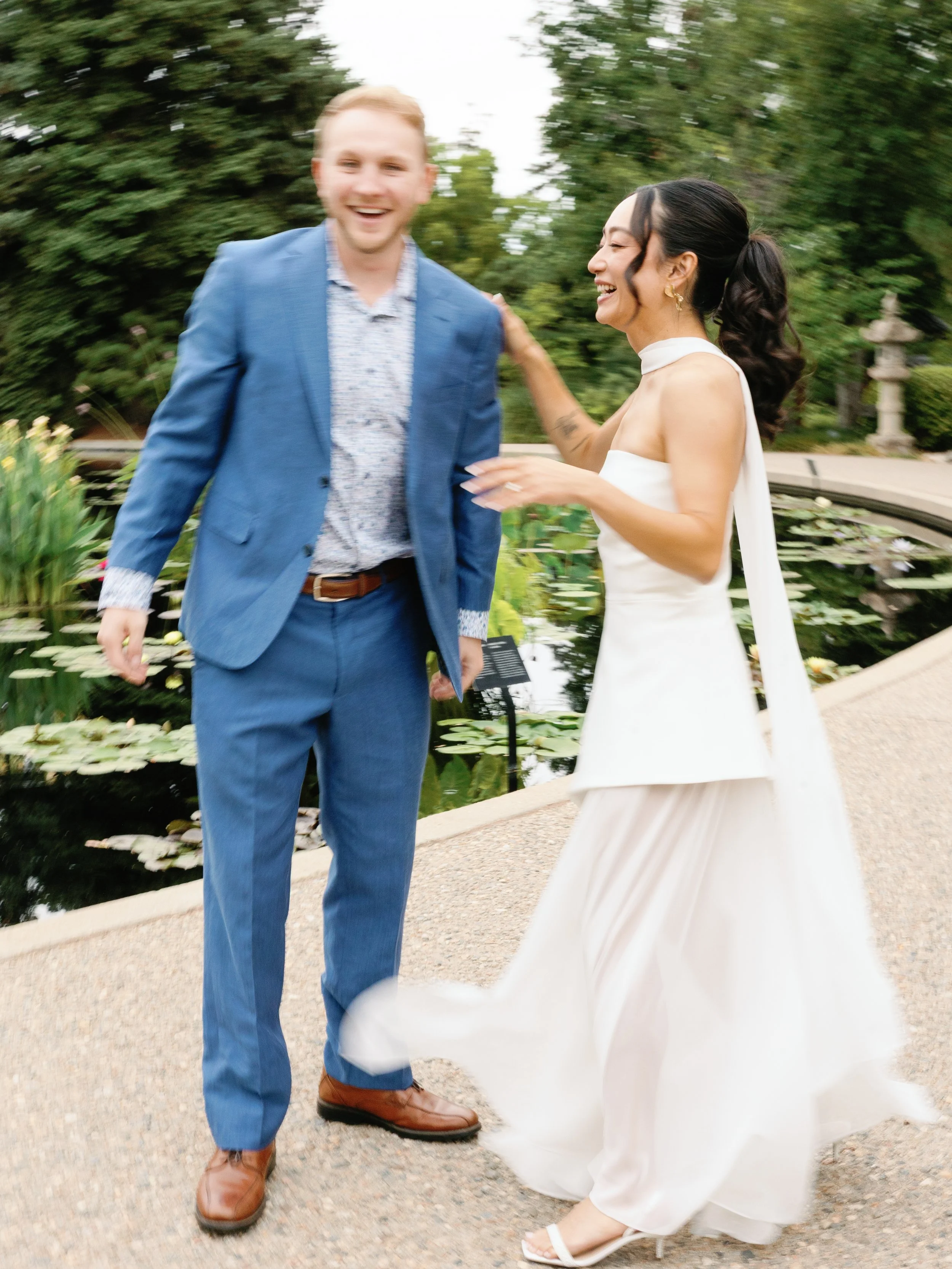 A joyful couple, a man in a blue suit and a woman in a white dress, laughing together outdoors near a pond with lily pads and greenery.