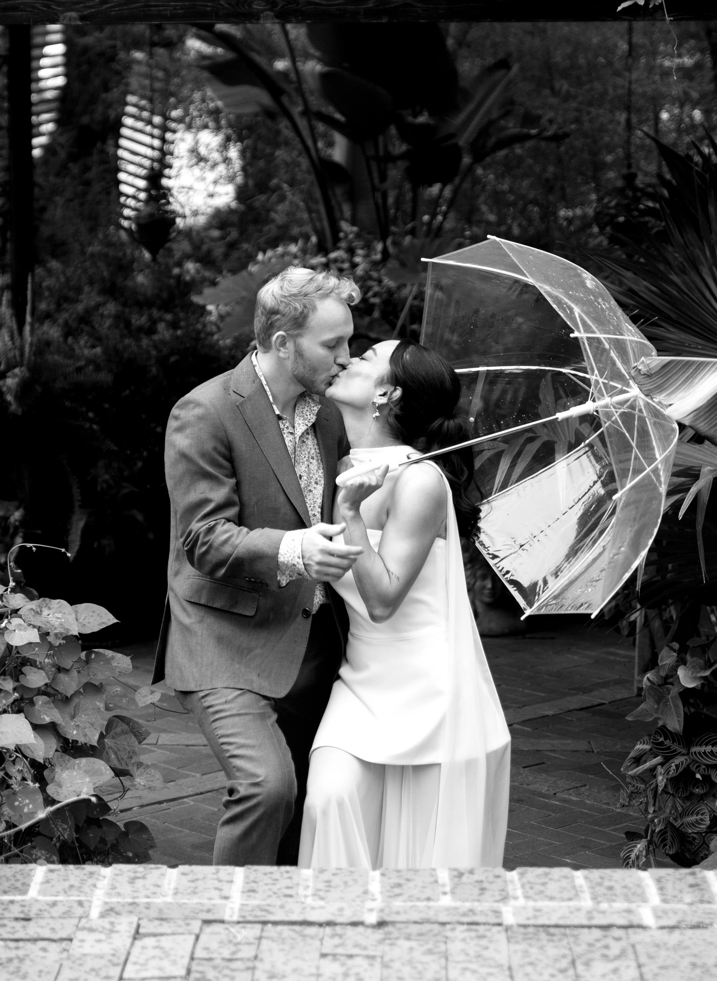 A couple sharing a kiss under a transparent umbrella in a lush outdoor setting.