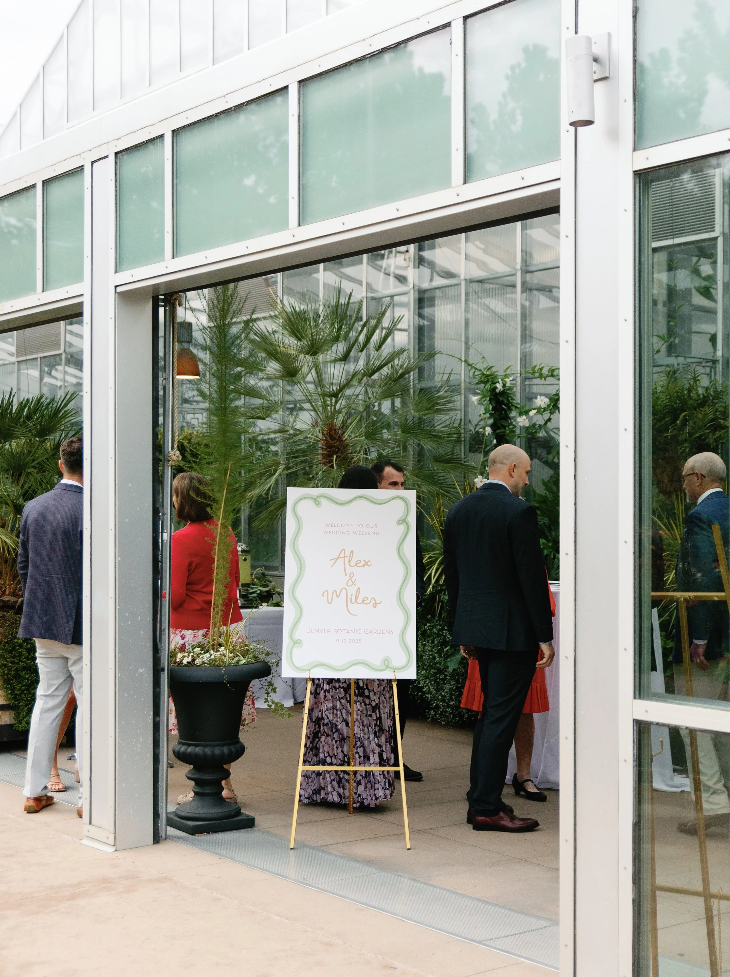 Guests at a wedding reception inside a greenhouse at Denver Botanic Gardens with a welcoming sign for Alex and Miles on September 12, 2023.