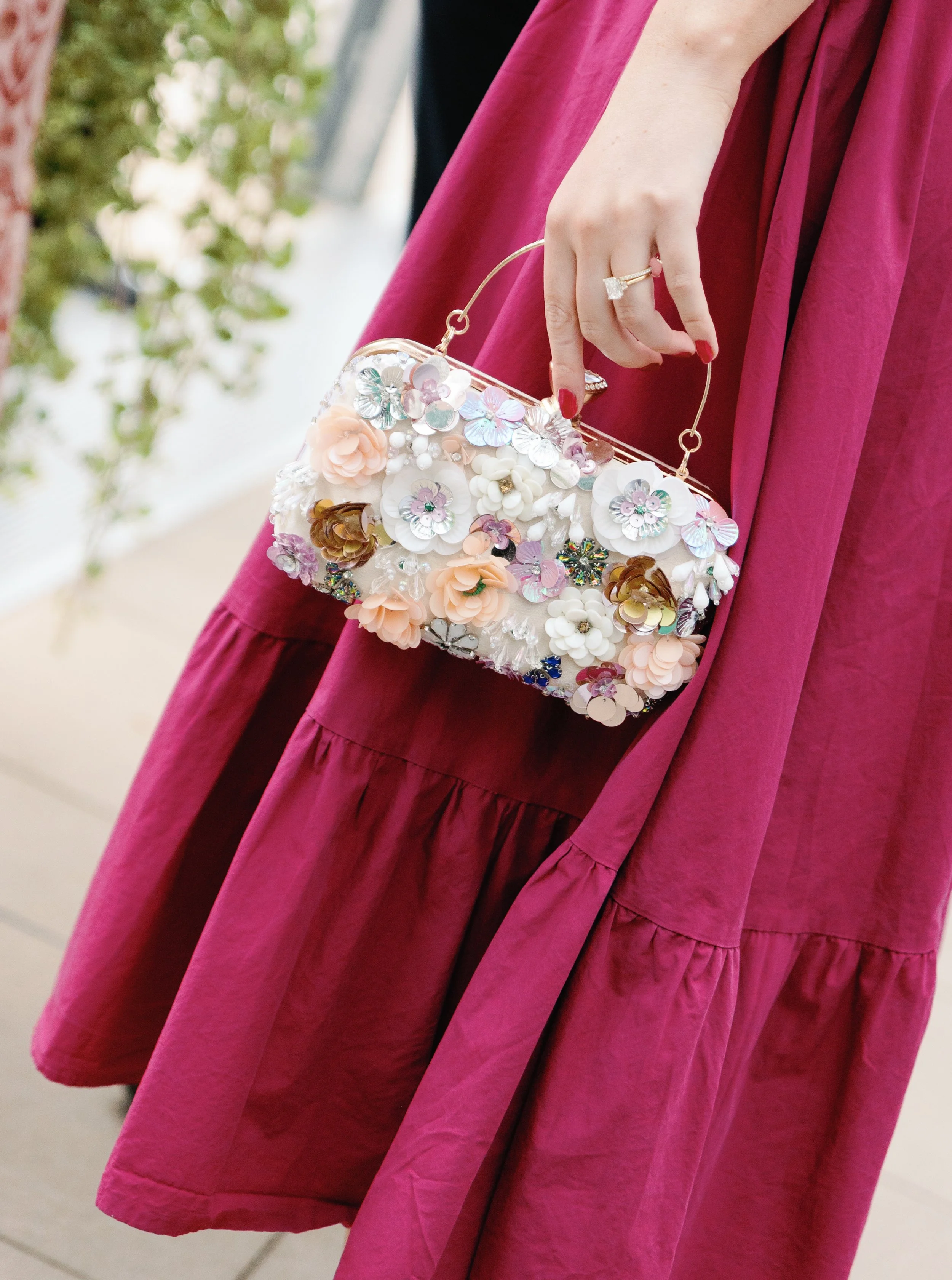 A woman wearing a burgundy dress holds a floral clutch purse with a gold handle and a ring on her finger, in an outdoor setting.