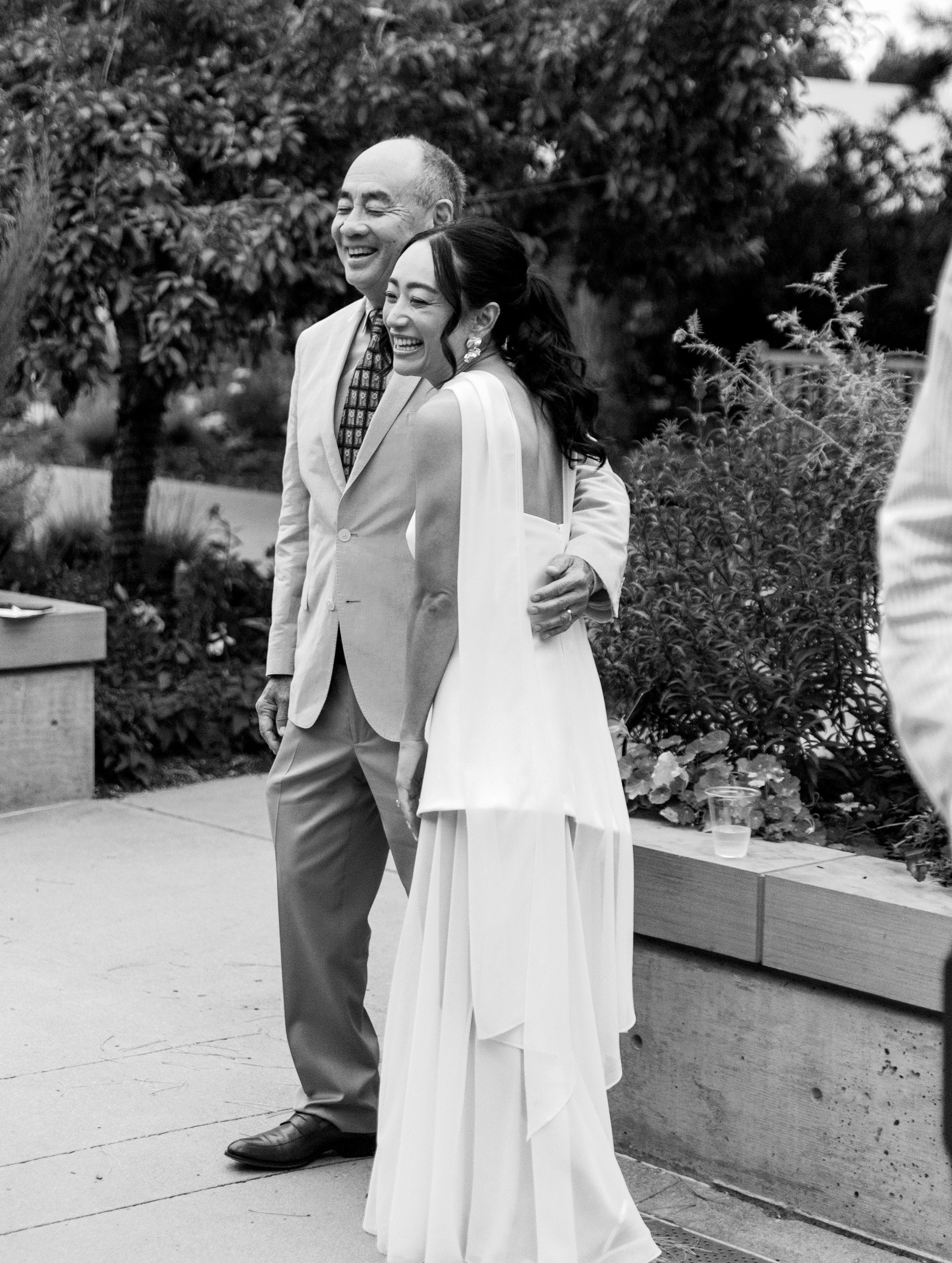 Black and white photo of a joyful couple at a wedding, standing outdoors with trees and plants in the background. The man is wearing a suit, and the woman is in a wedding dress, both smiling.