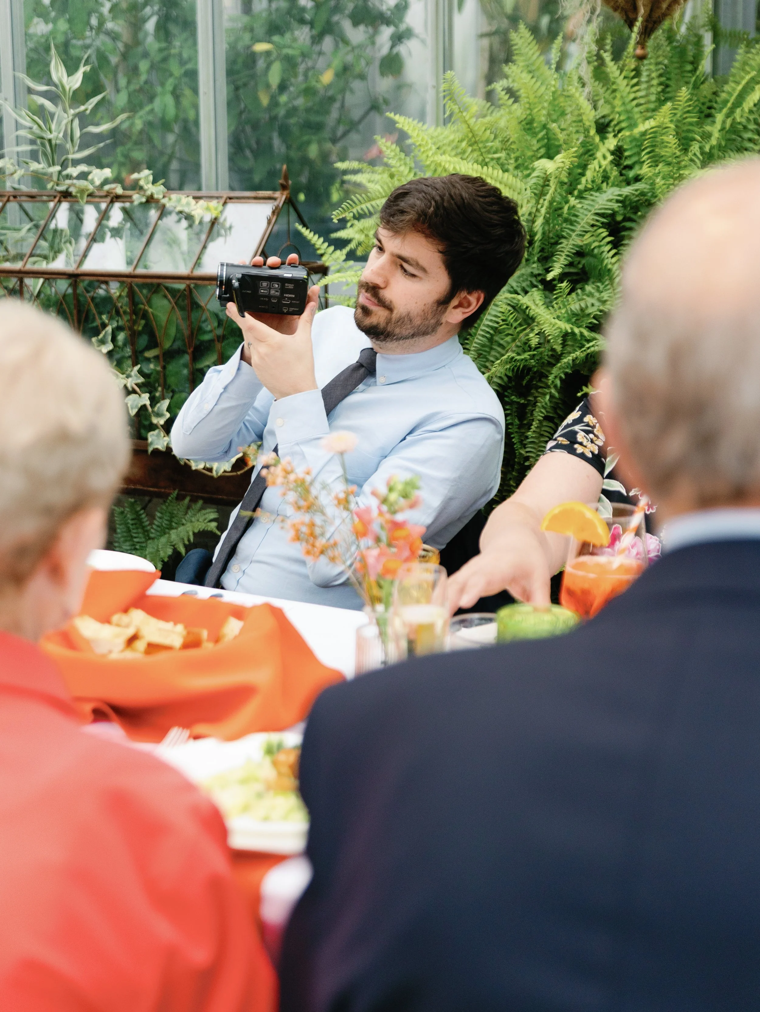 A man with dark hair and a beard is sitting at a table, holding a camera and looking at it. The table is set with food, drinks, and a small bouquet of flowers. There are several older people sitting at the table, and the setting appears to be a green