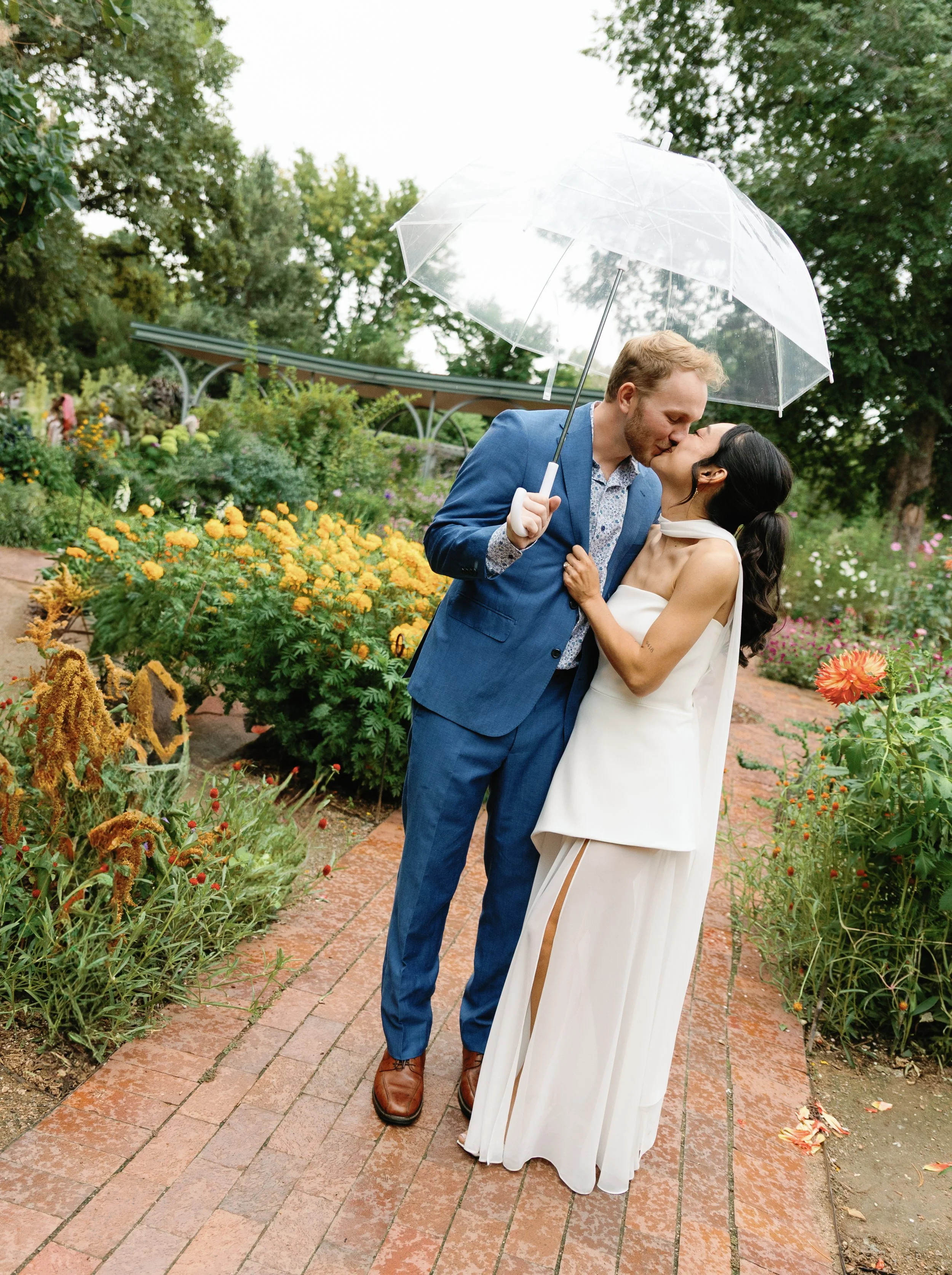 A couple in wedding attire sharing a kiss under a clear umbrella in a garden with colorful flowers.