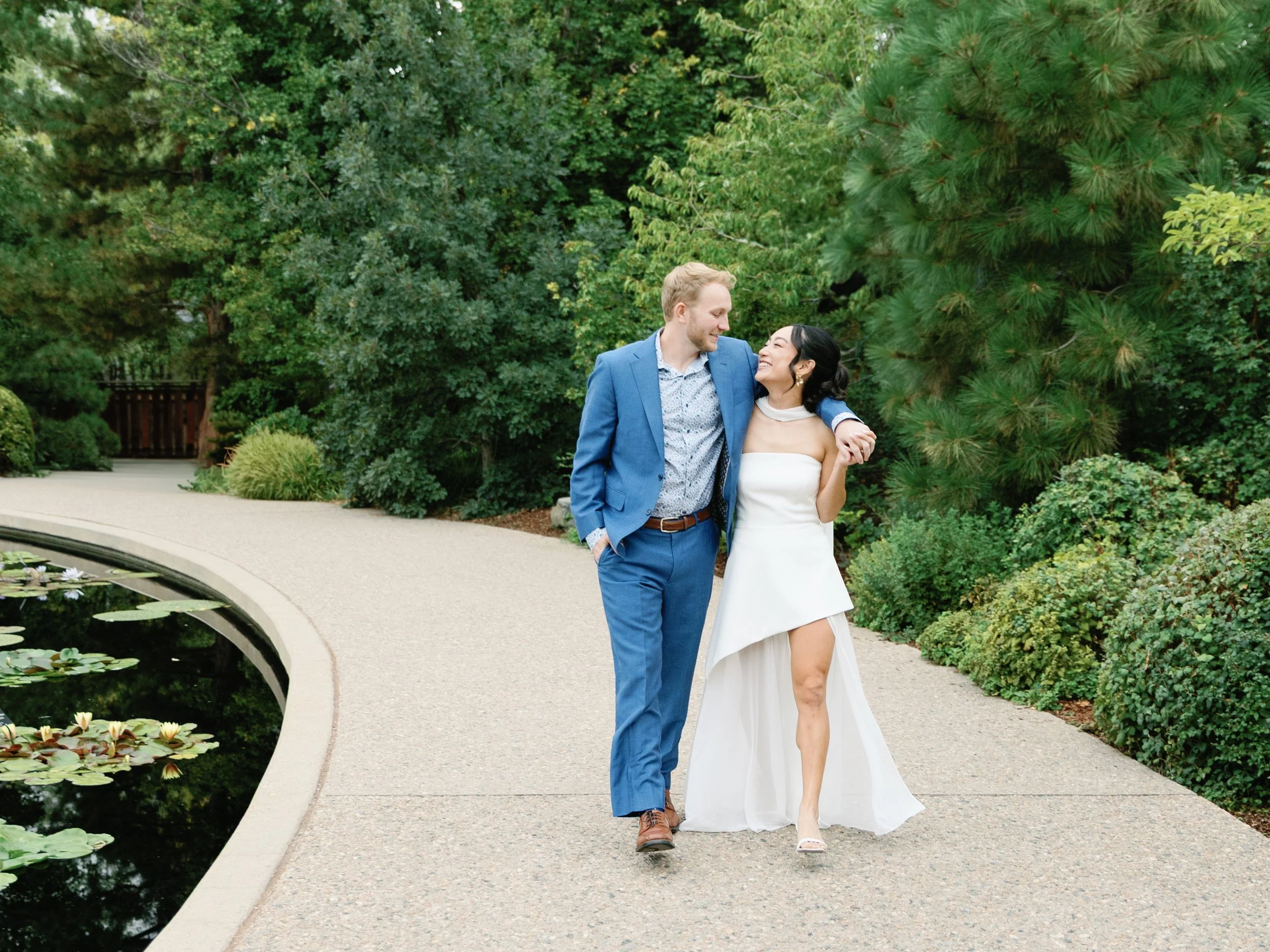 A couple walking arm in arm on a garden path, smiling and looking at each other, with trees and a pond with lily pads nearby.