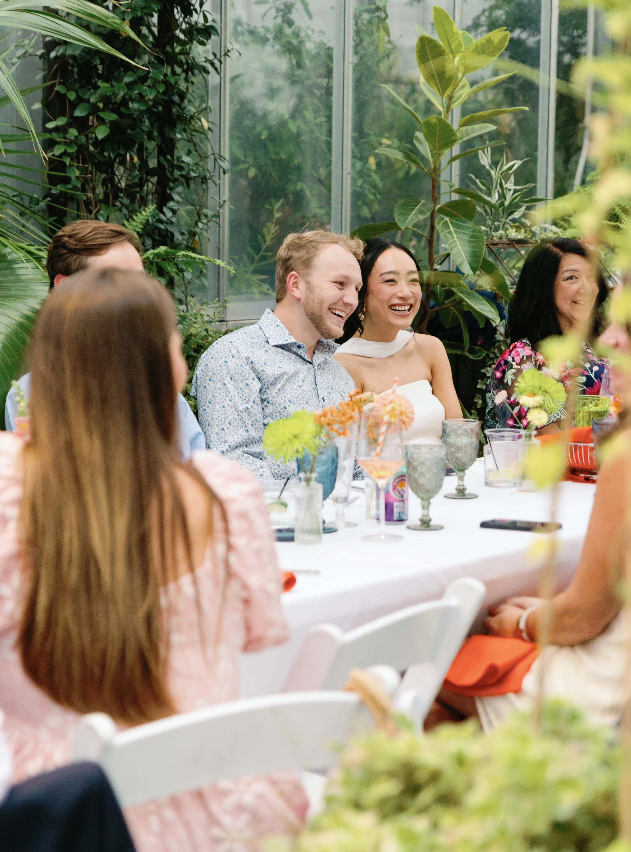People sitting at a table in a greenhouse filled with lush green plants, laughing and enjoying a social gathering.