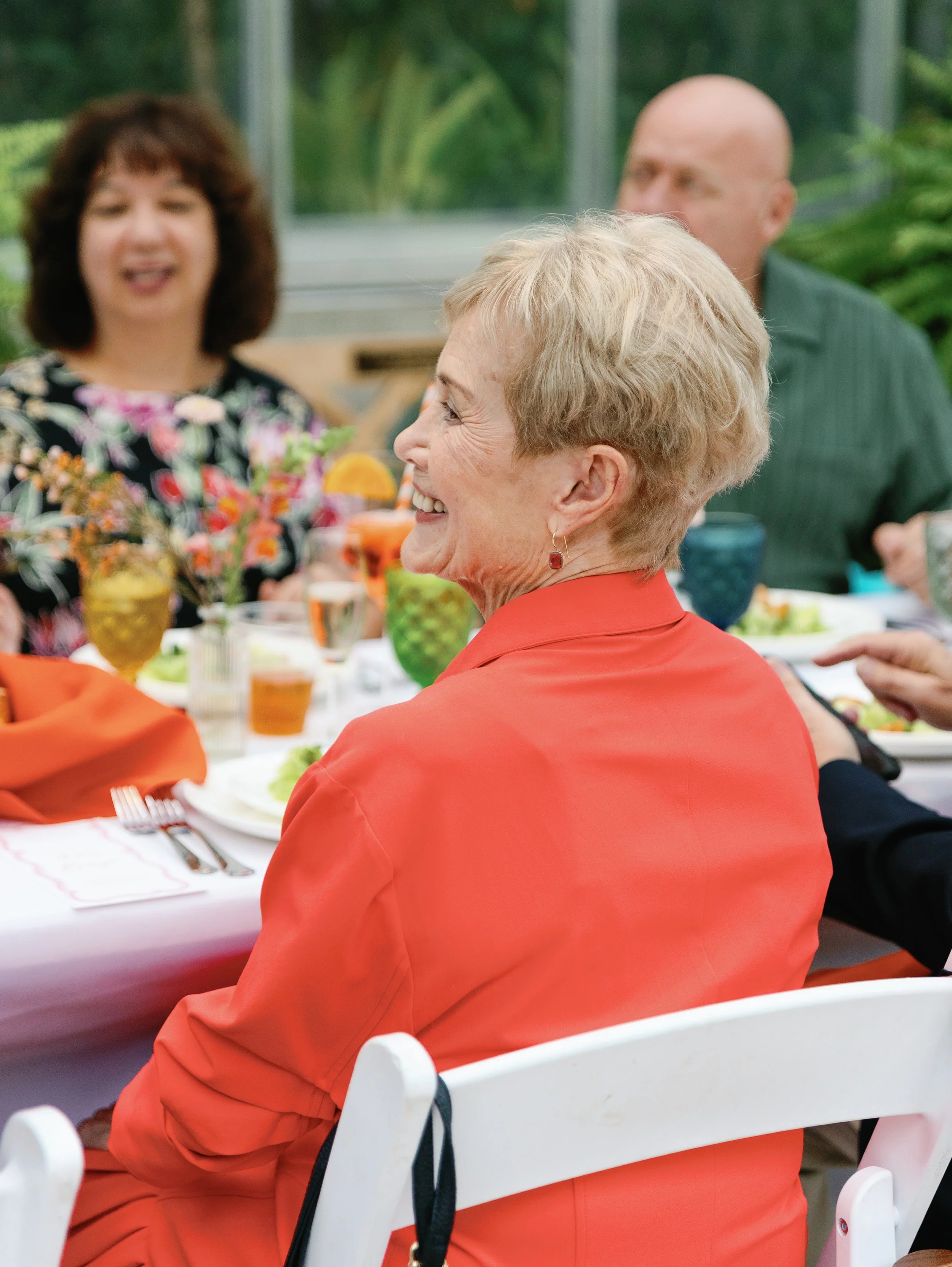 Smiling elderly woman in a red jacket sitting at a dinner table with other people, enjoying a social gathering outdoors.