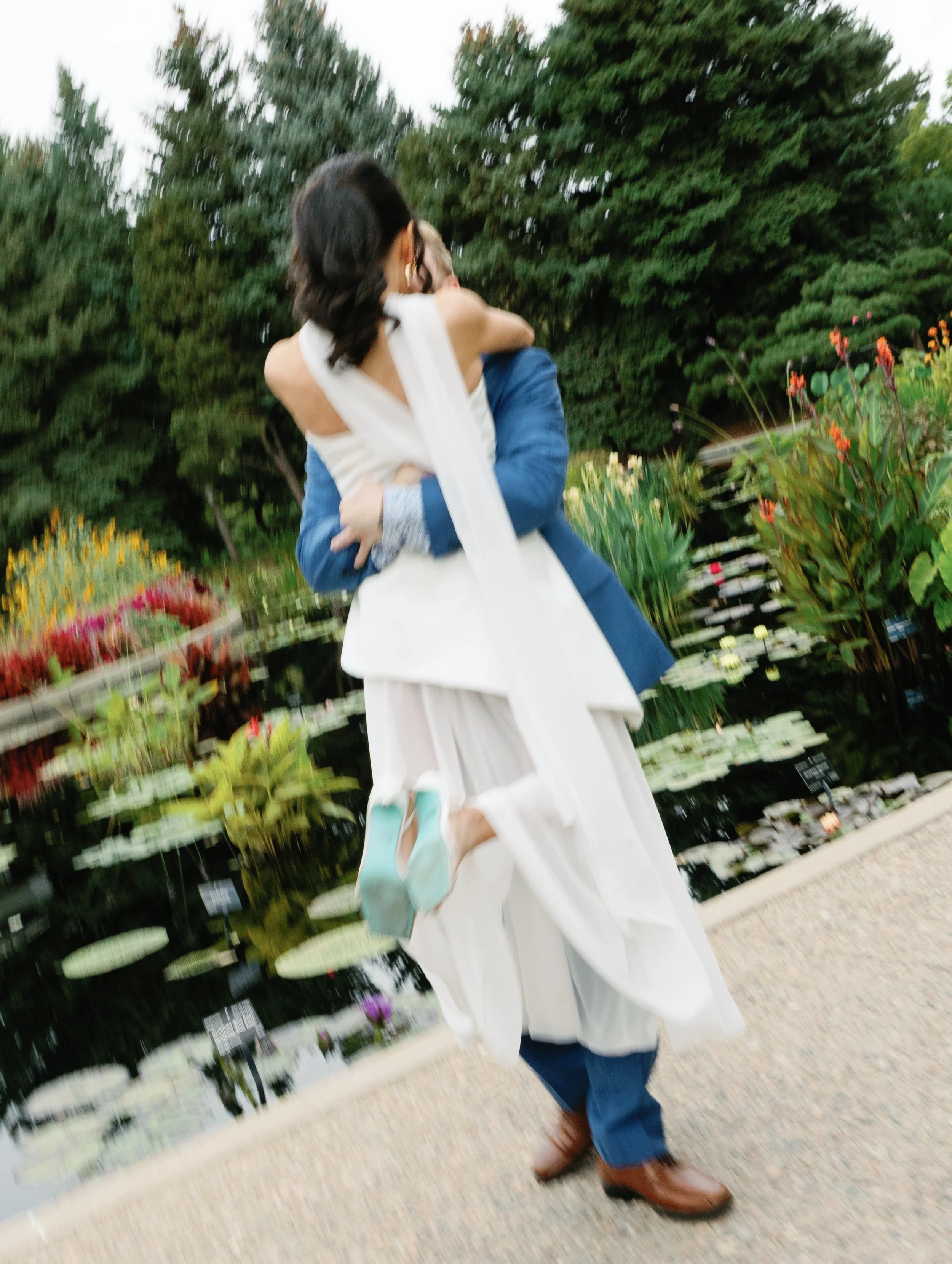 A couple is dancing by a pond with lily pads and colorful flowers in the background.