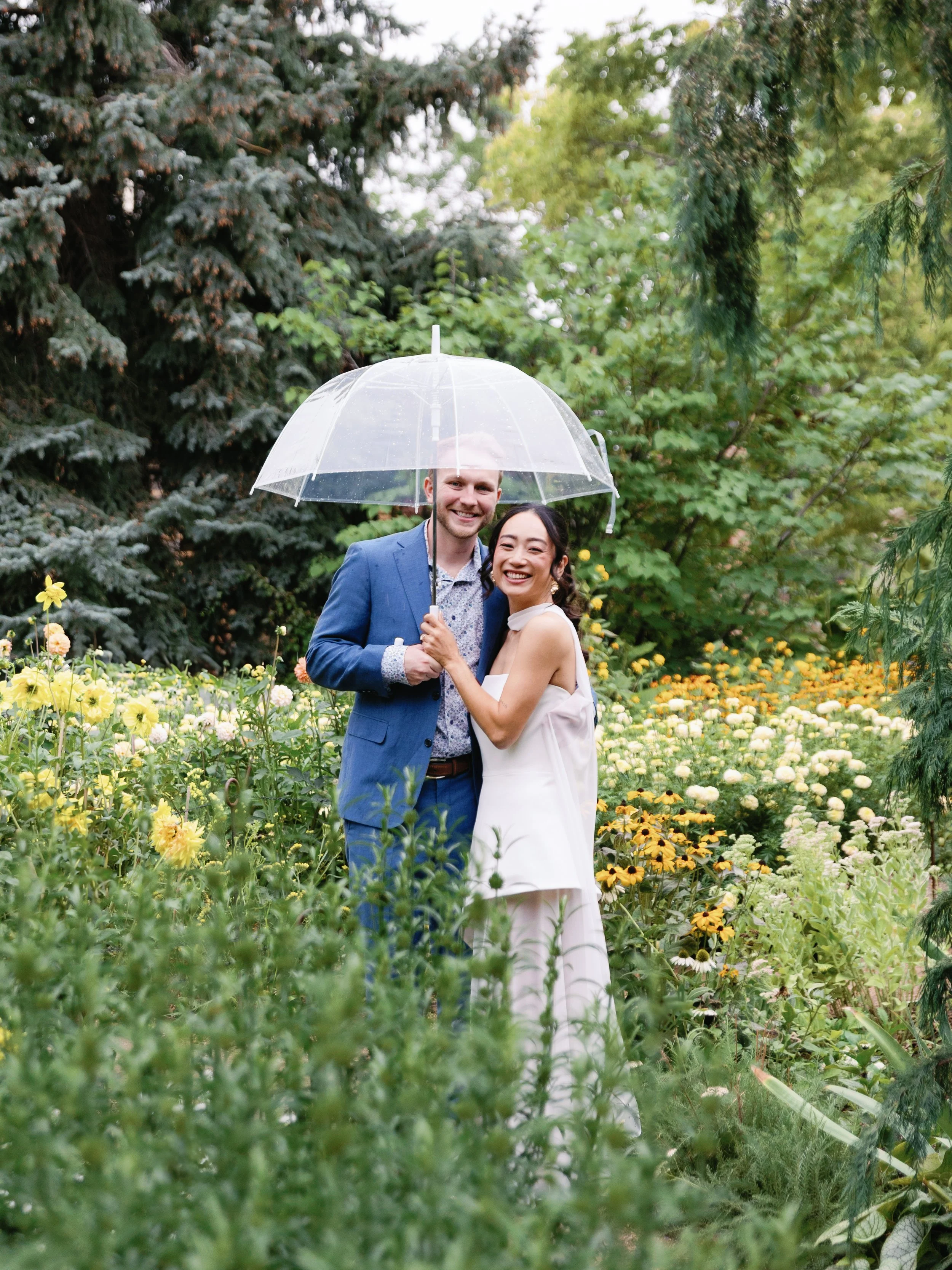 A happy couple stands in a garden surrounded by yellow and white flowers, sharing an umbrella on a rainy day, smiling at the camera.