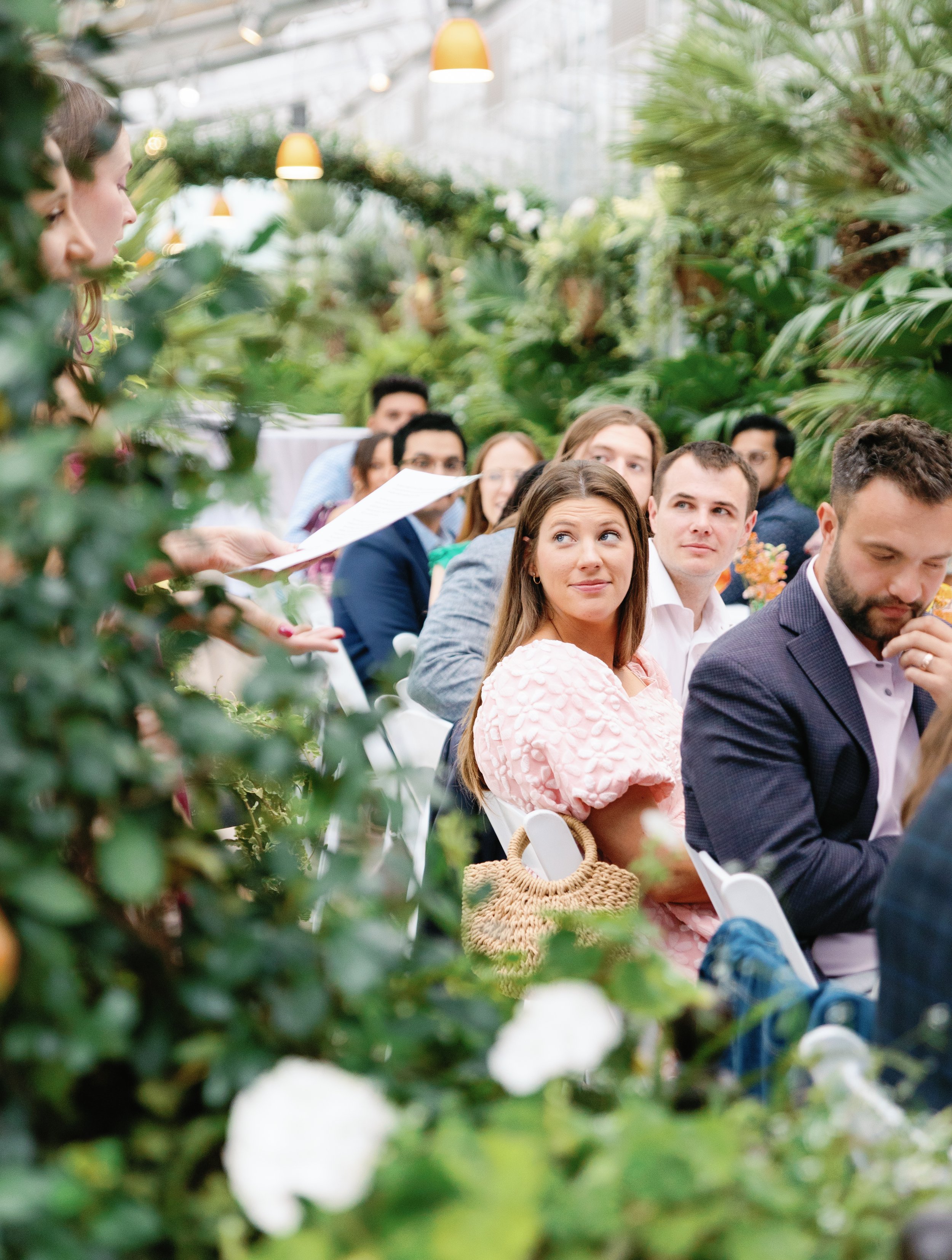 A group of people seated in a lush, green greenhouse, attentively listening to a woman speaking.