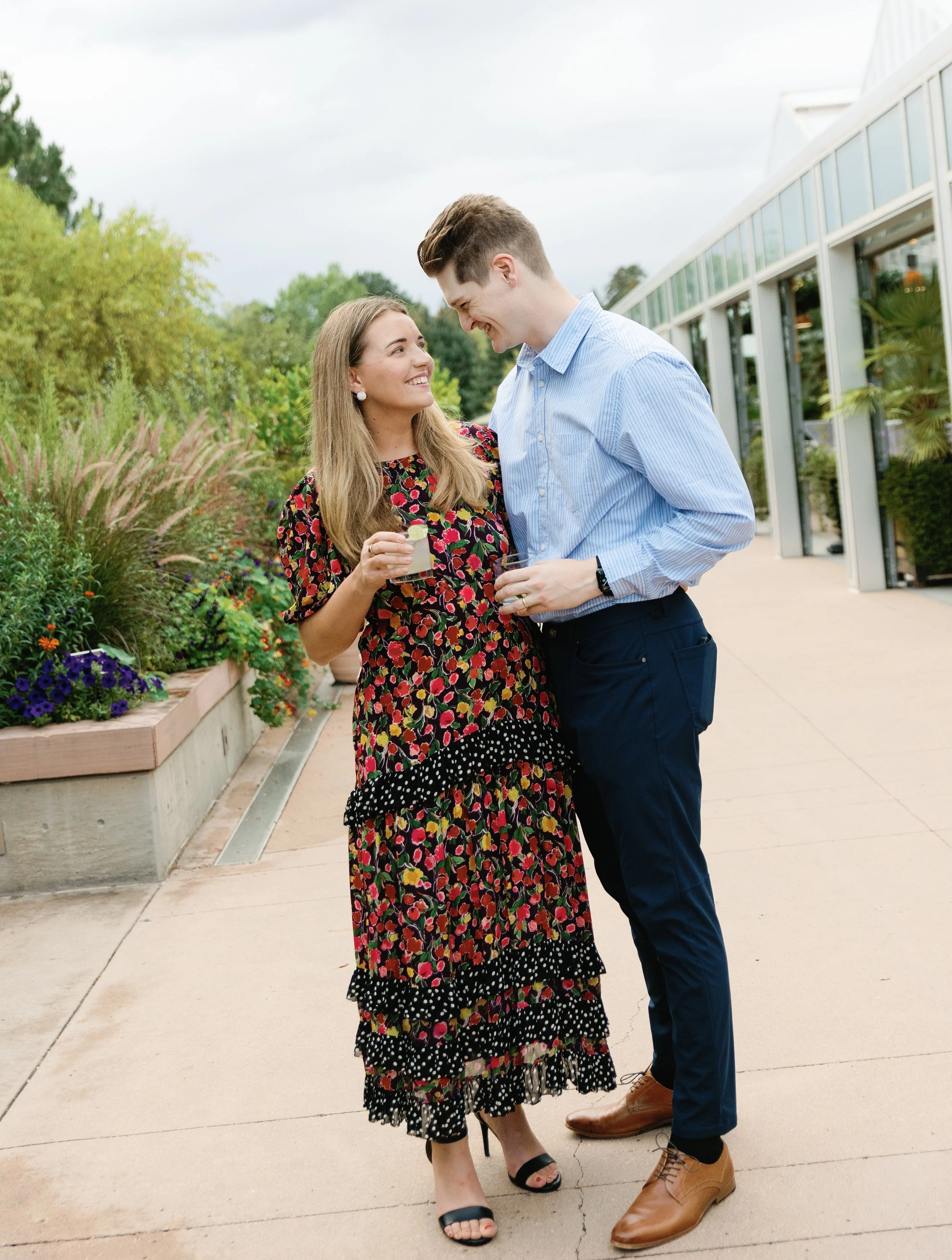 A man and woman talking and smiling outside near a greenhouse and garden, holding drinks, with trees and a cloudy sky in the background.
