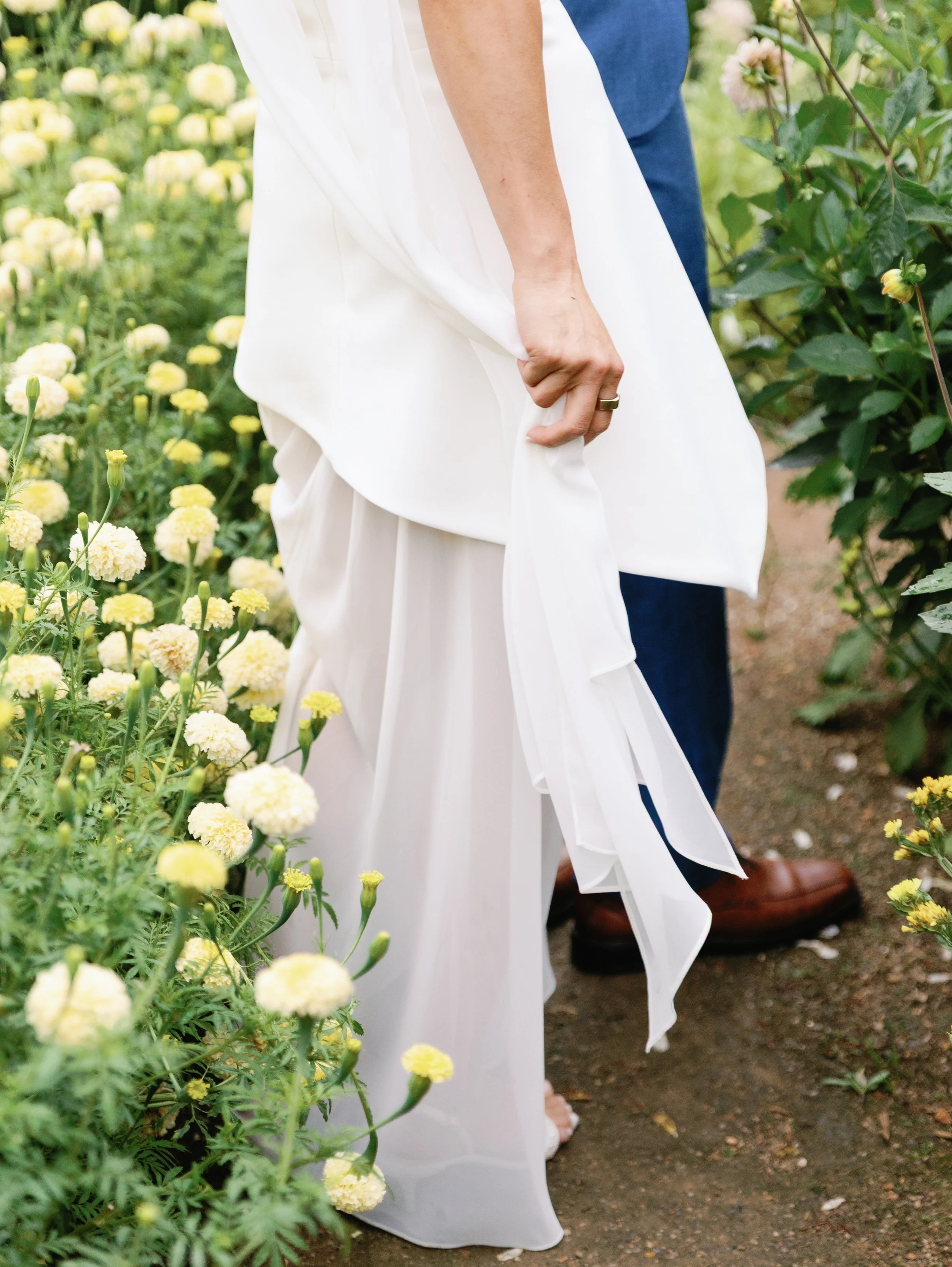 A bride in a white wedding dress holding the fabric of her dress, standing among yellow and white flowers while a groom in blue pants and brown shoes stands behind her.