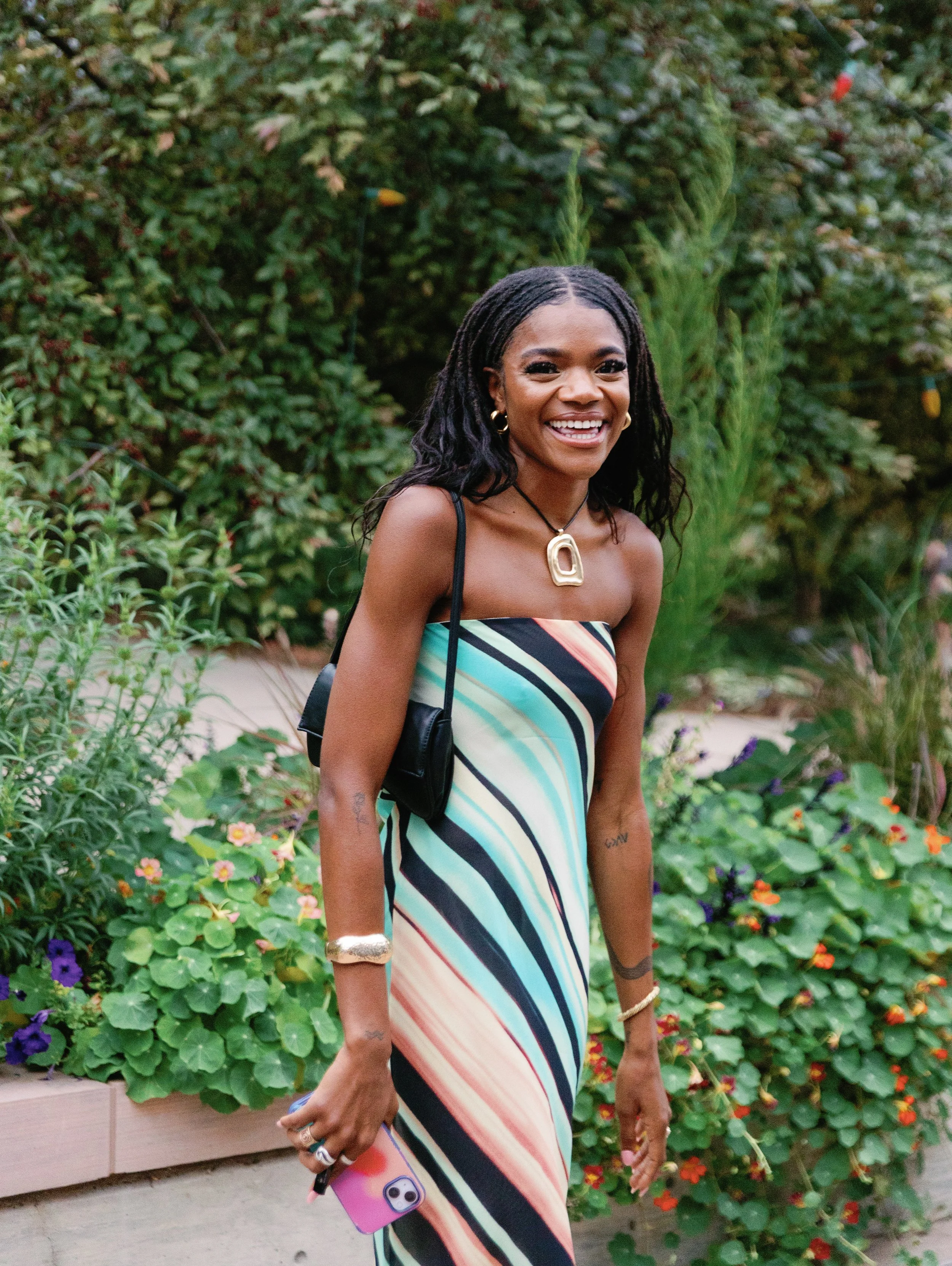 A woman with dark curly hair is smiling and standing outdoors in front of green plants and flowers. She is wearing a colorful striped strapless dress, jewelry, and carrying a black purse and a pink phone.