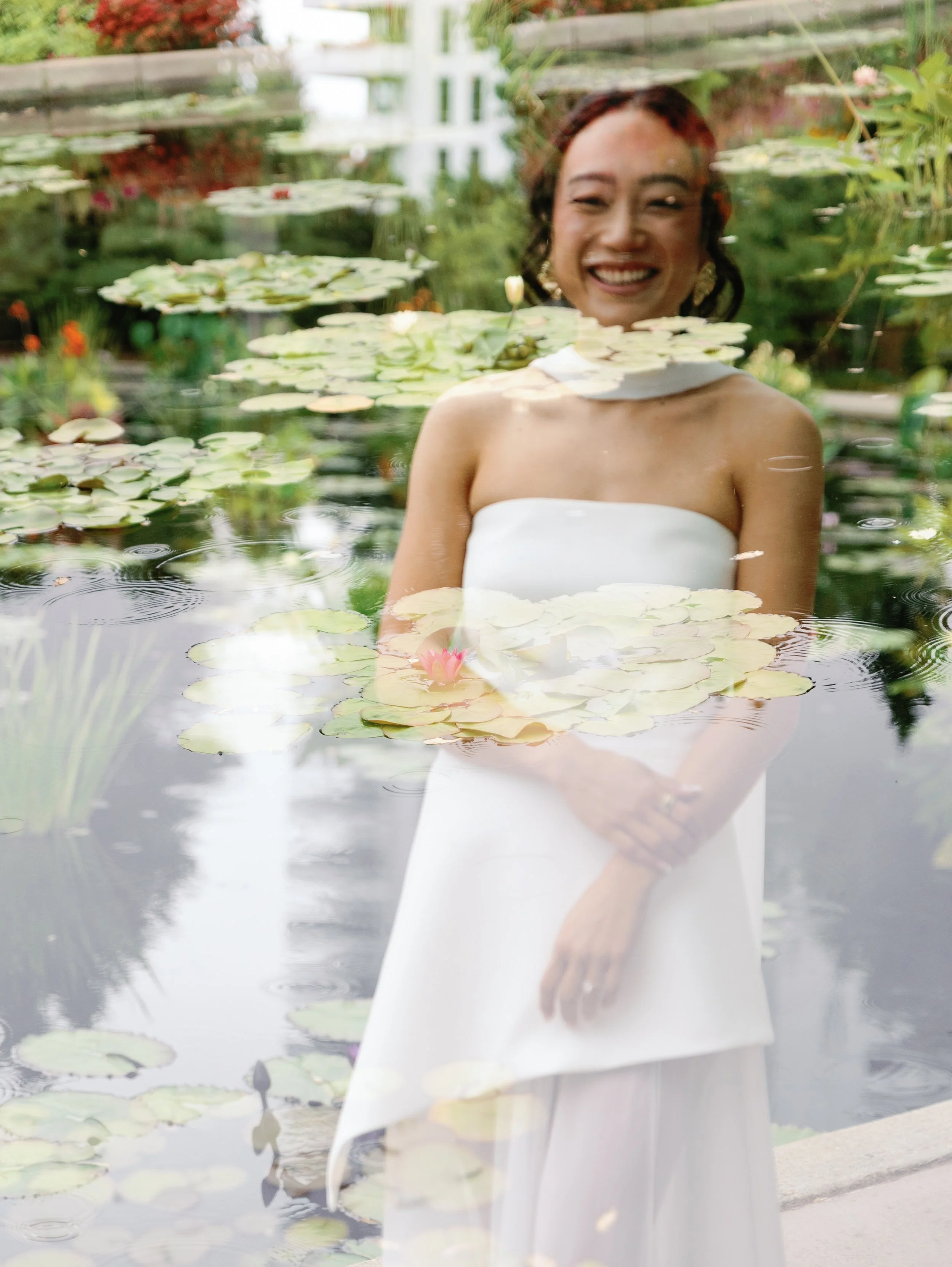 A woman in a white strapless dress smiling through a glass pond with lily pads and blooming flowers.