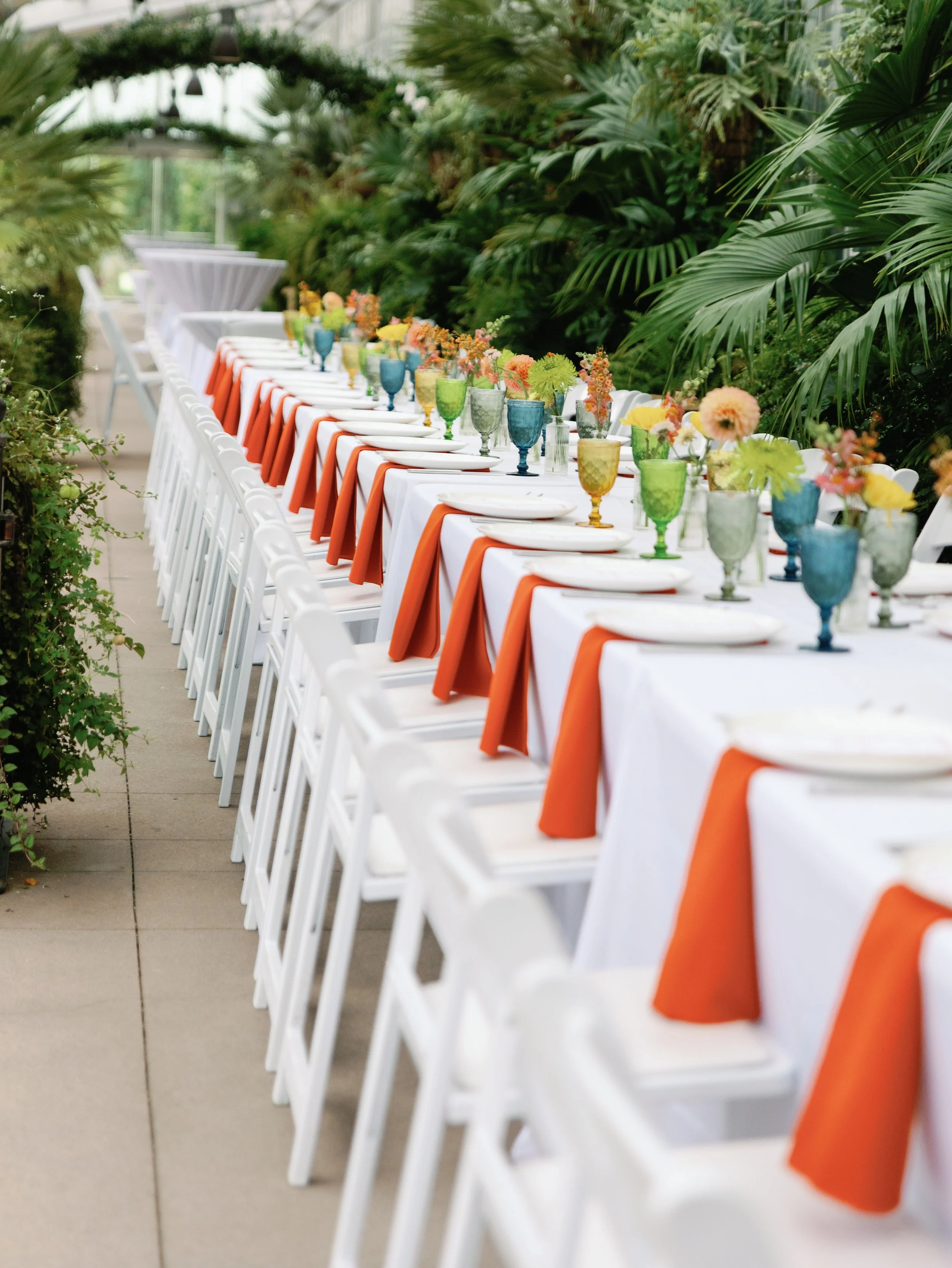 Long dining table covered with white tablecloth, orange napkins, colorful glassware, and floral centerpieces, set outdoors in a lush green garden.