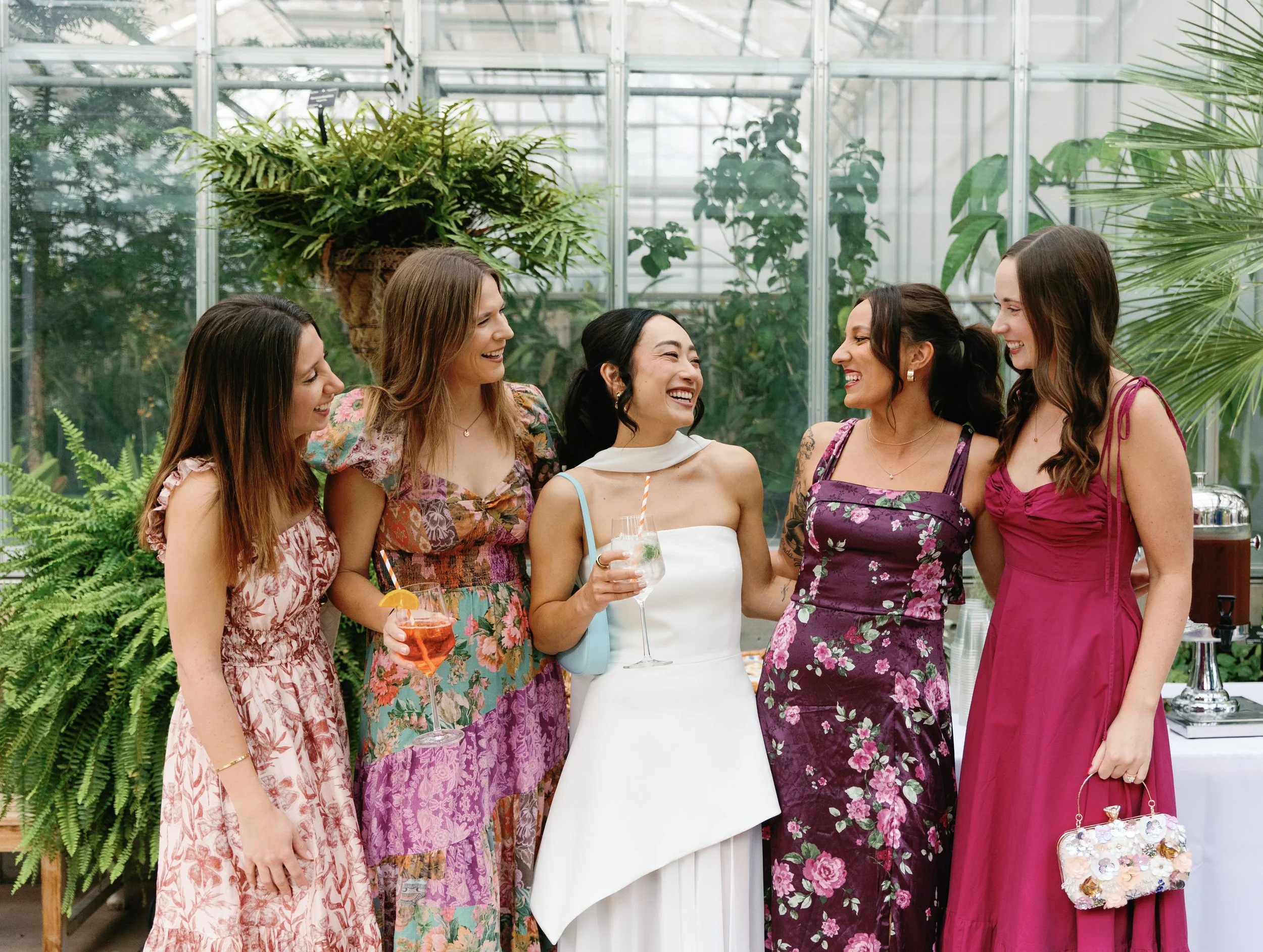 Group of five women dressed in colorful dresses, smiling, and enjoying drinks in a greenhouse with lush green plants.