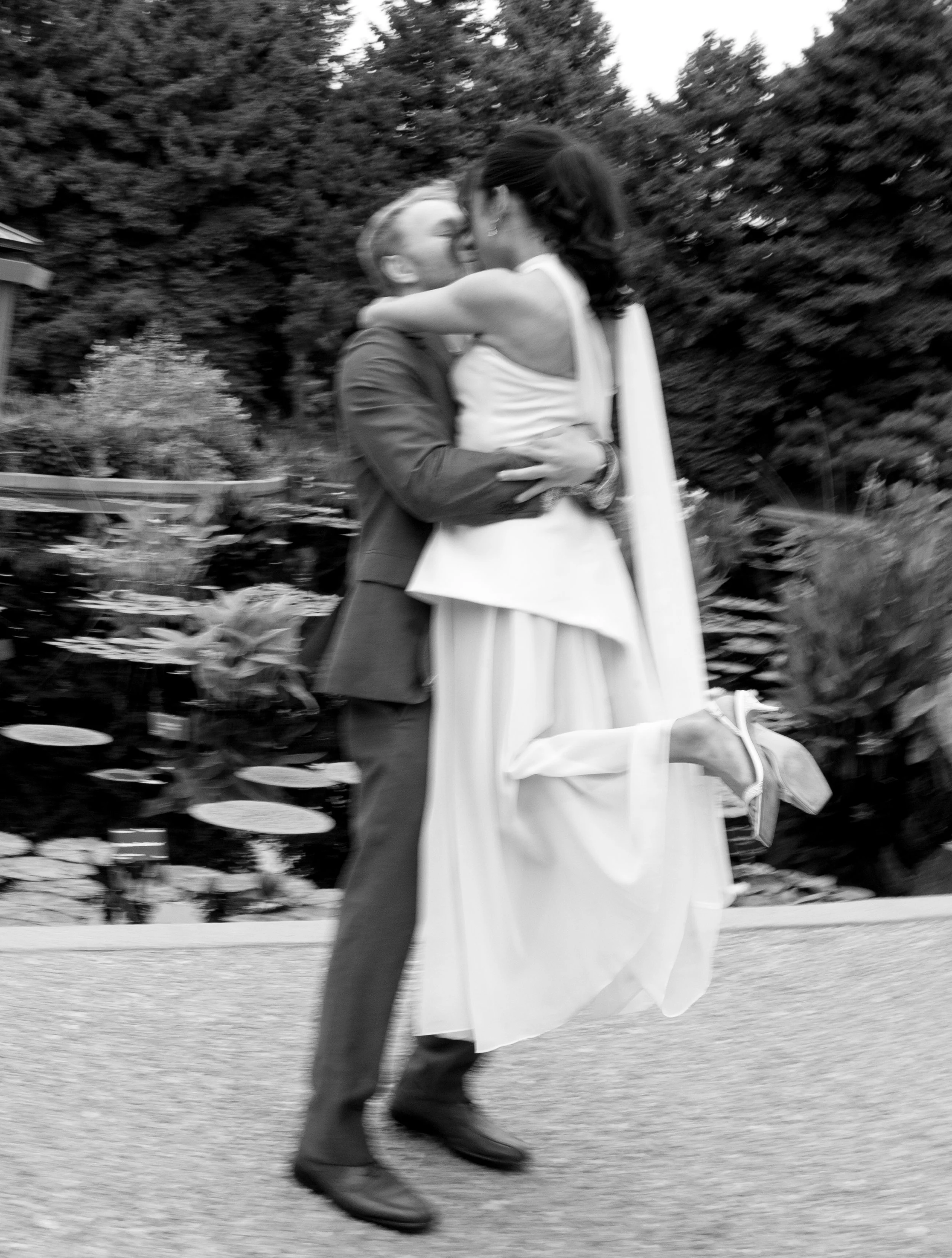 A black and white photo of a man lifting a woman in a wedding dress, both smiling and embracing in a garden with trees and water lilies.
