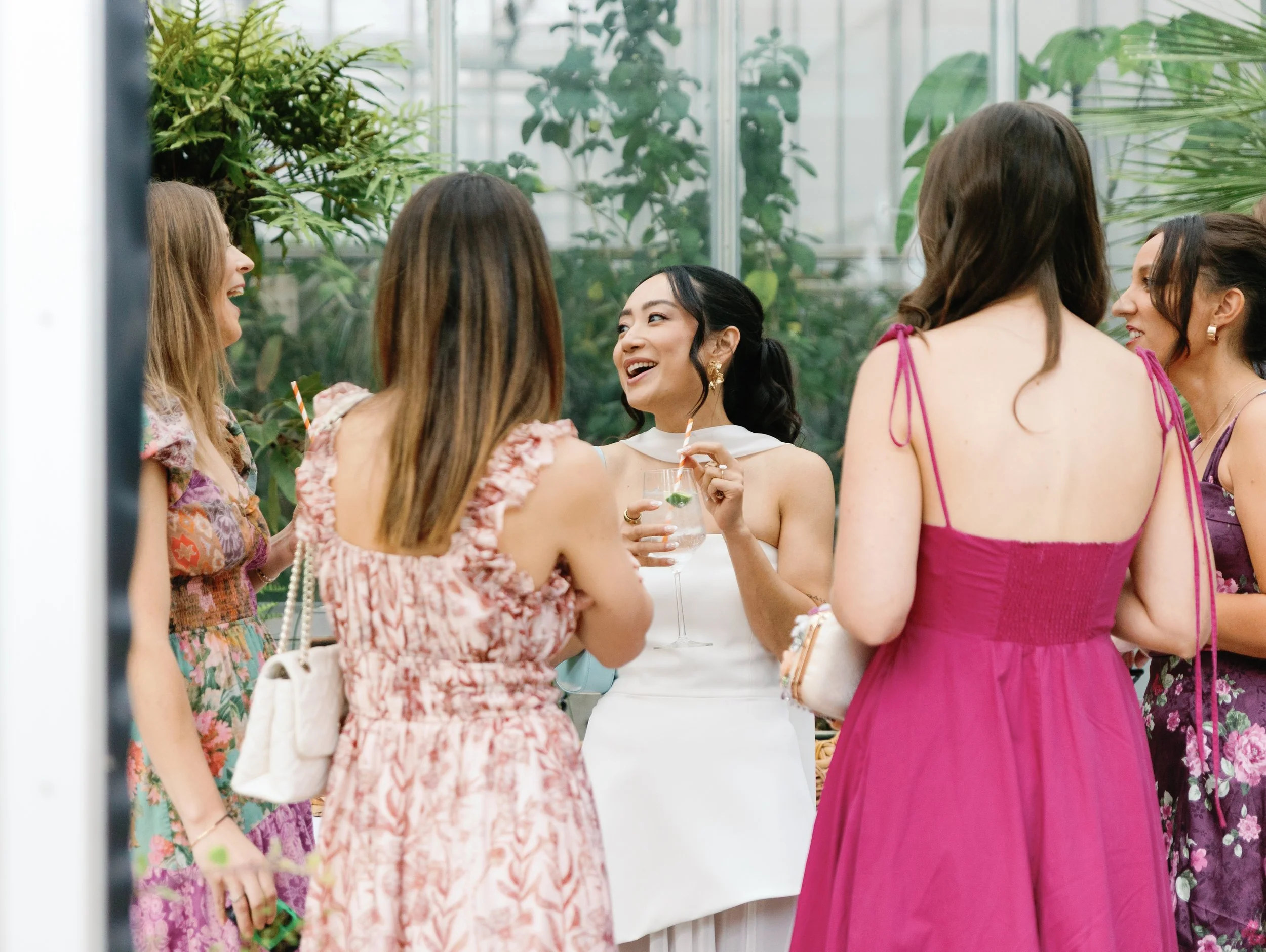 Group of women at a social gathering in a greenhouse, chatting and smiling, one holding a glass of cocktail with a straw and mint garnish.