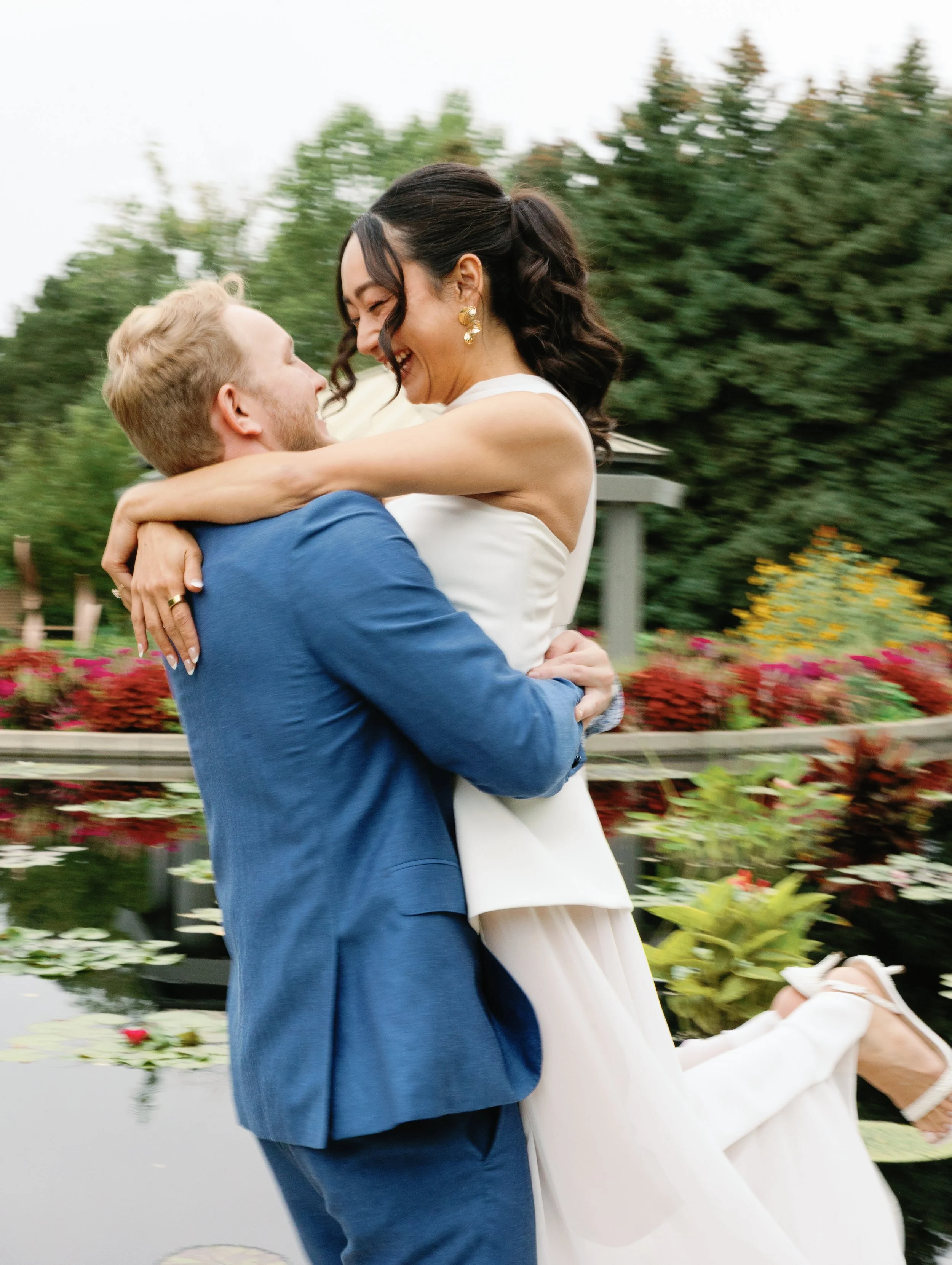 A happy couple is embracing outdoors near a pond, with colorful flowers and greenery in the background, during daytime.