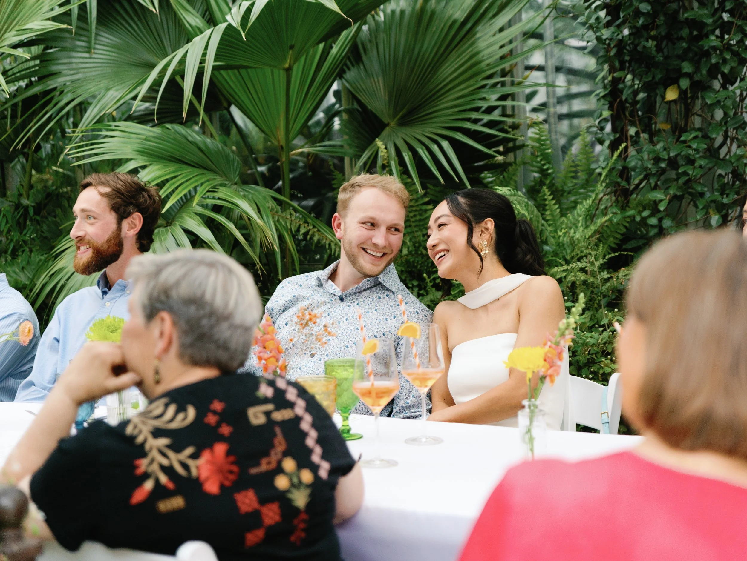 People enjoying a garden party or celebration, seated at a table with drinks and flowers, surrounded by lush greenery.