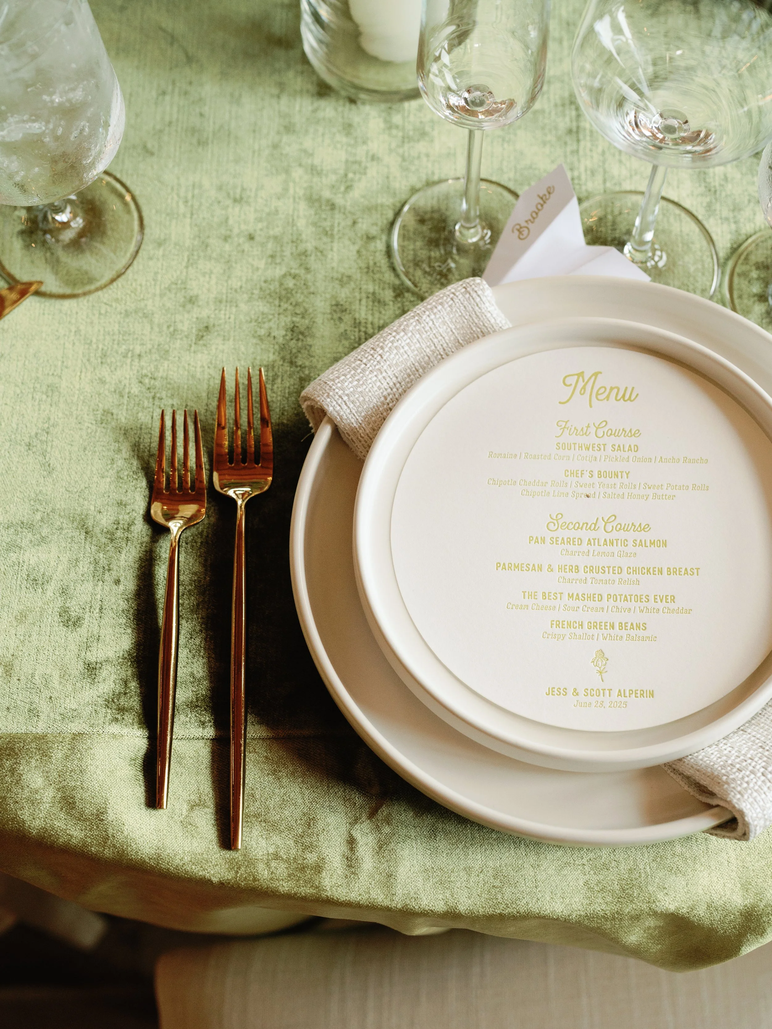 A place setting at a formal event with a green tablecloth, two gold forks, a white napkin, a white dinner plate with a printed menu, and stemmed wine glasses in the background.