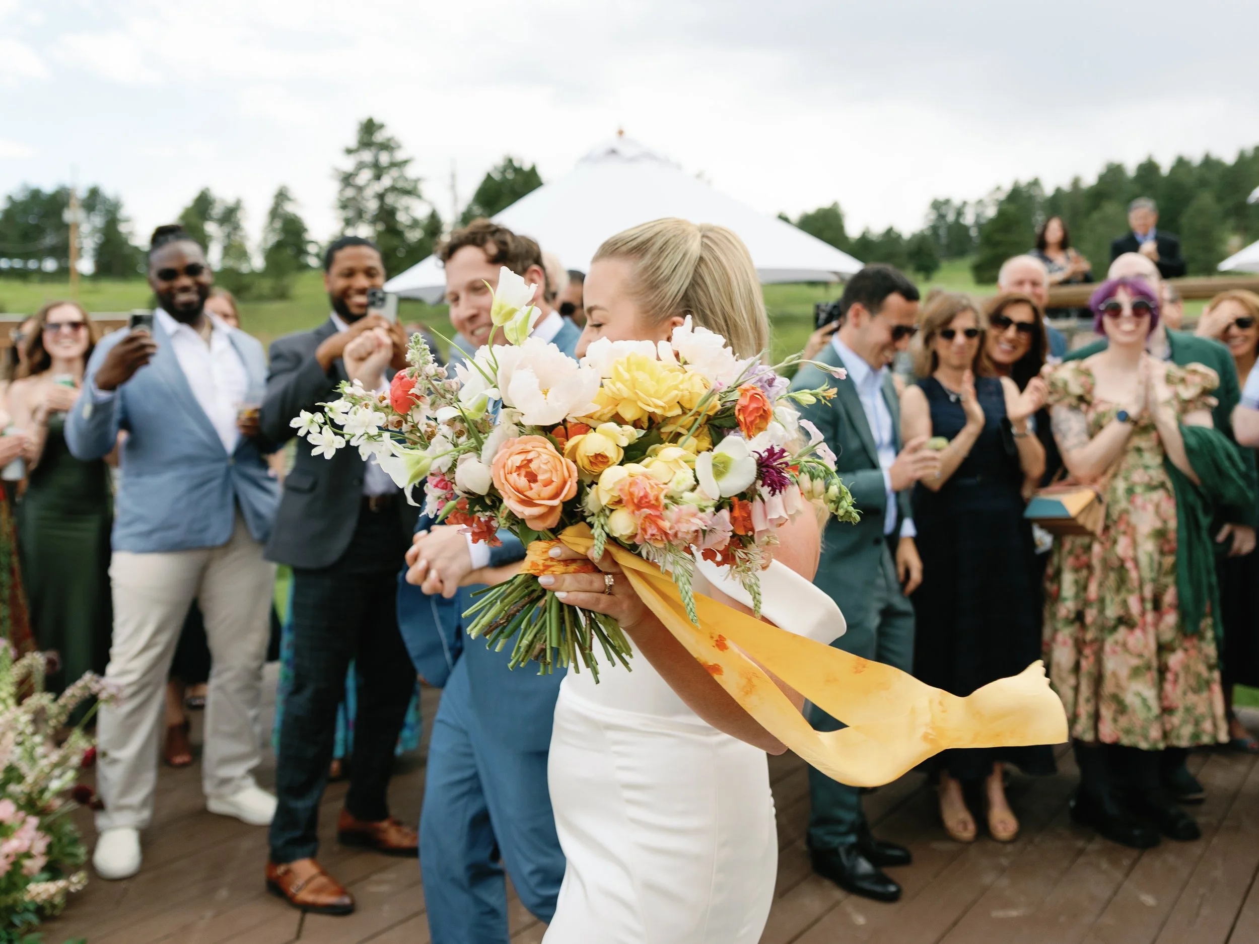 Bride holding a large bouquet of flowers with a ribbon, surrounded by wedding guests at an outdoor wedding reception.