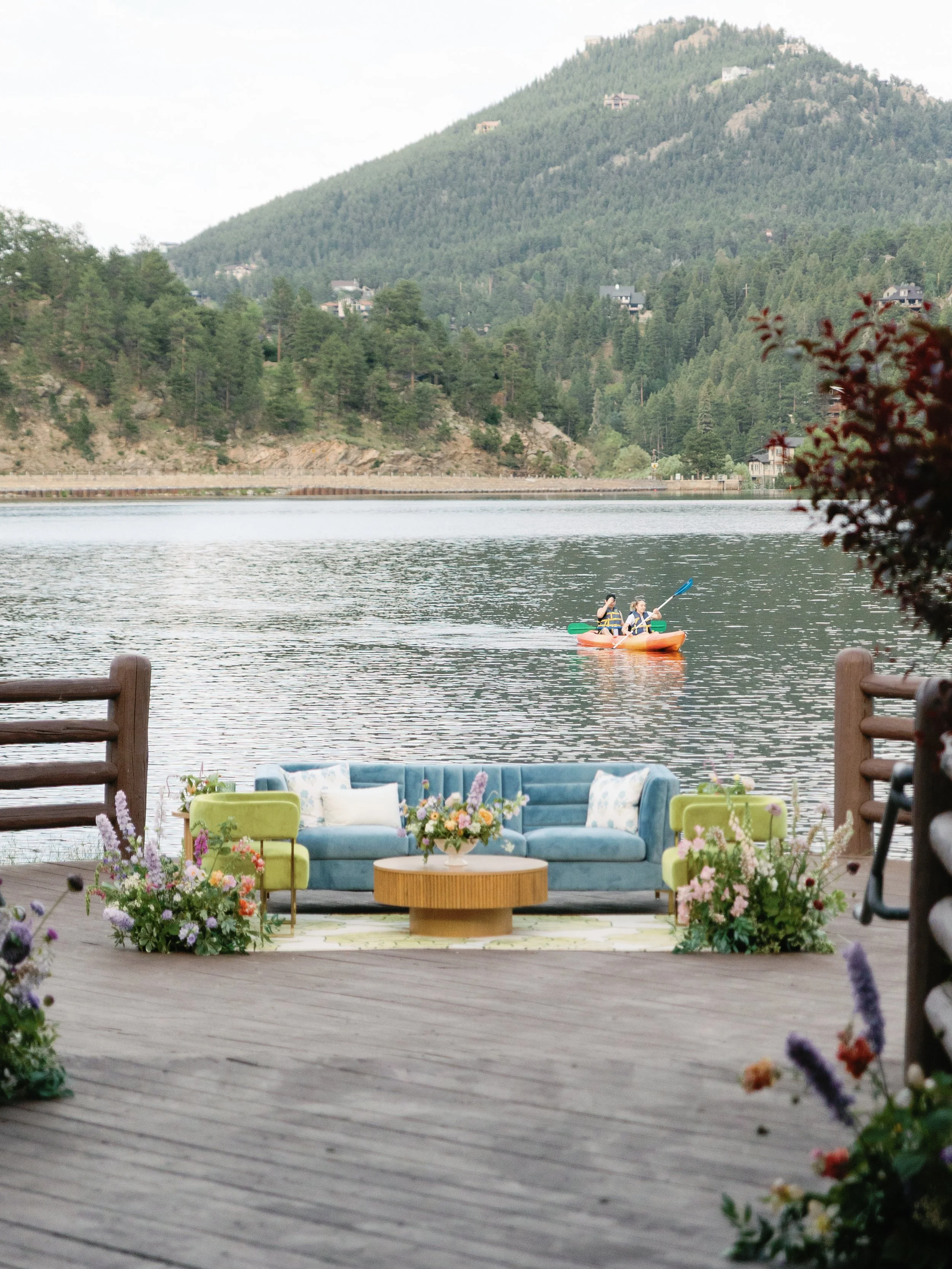A lakeside scene with a teal sofa and two matching chairs on a wooden deck surrounded by flowers, overlooking a lake with two people kayaking against a backdrop of green mountains.