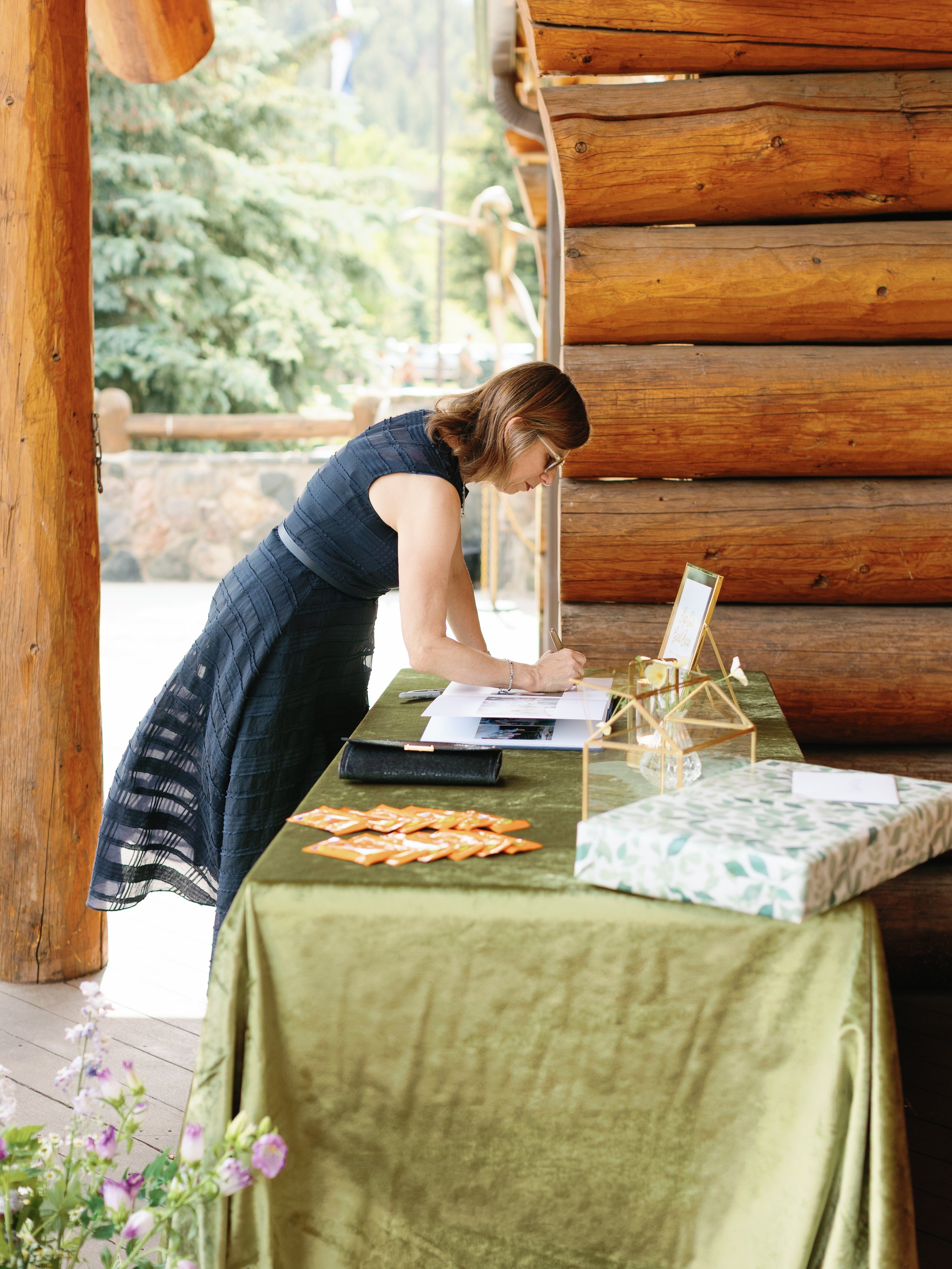Woman in a dark dress writing at a table covered with green fabric outside a log cabin.