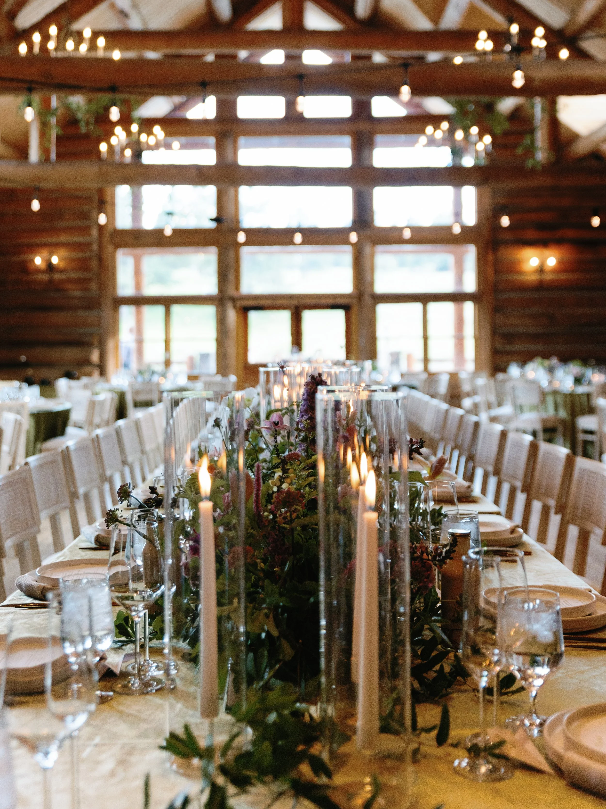 Wedding reception table with floral centerpiece, candles, and glassware in a rustic wooden barn with string lights.