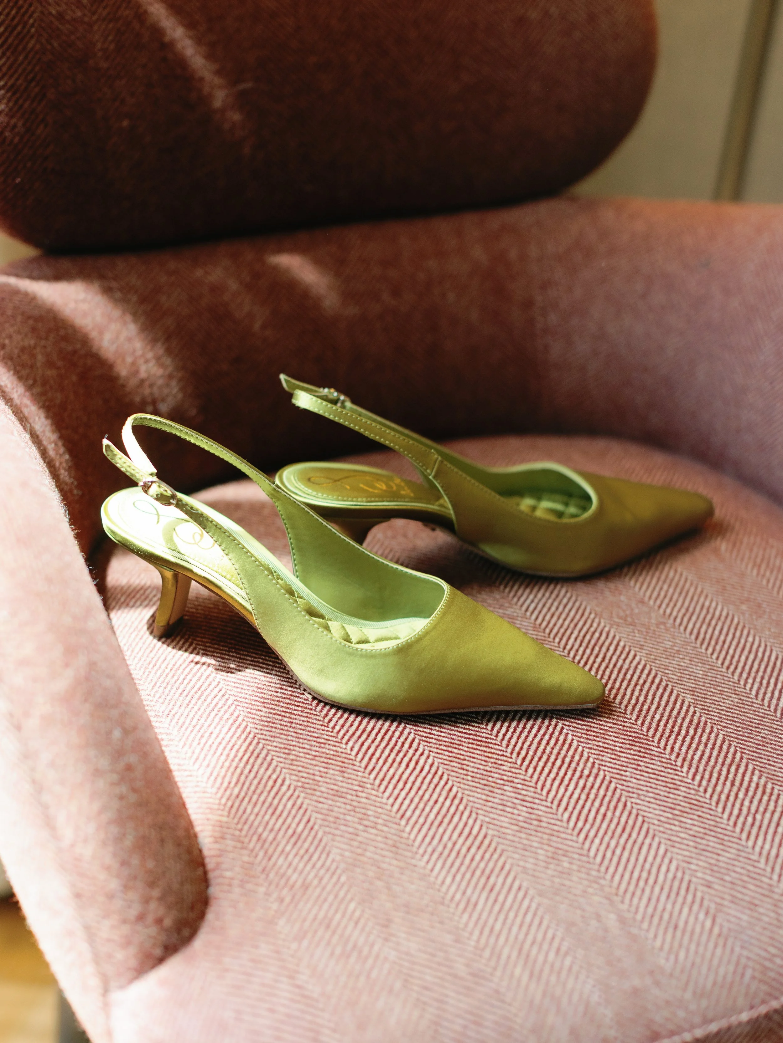 A pair of green satin pointed-toe slingback heels with a small heel, placed on a pinkish-red armchair.