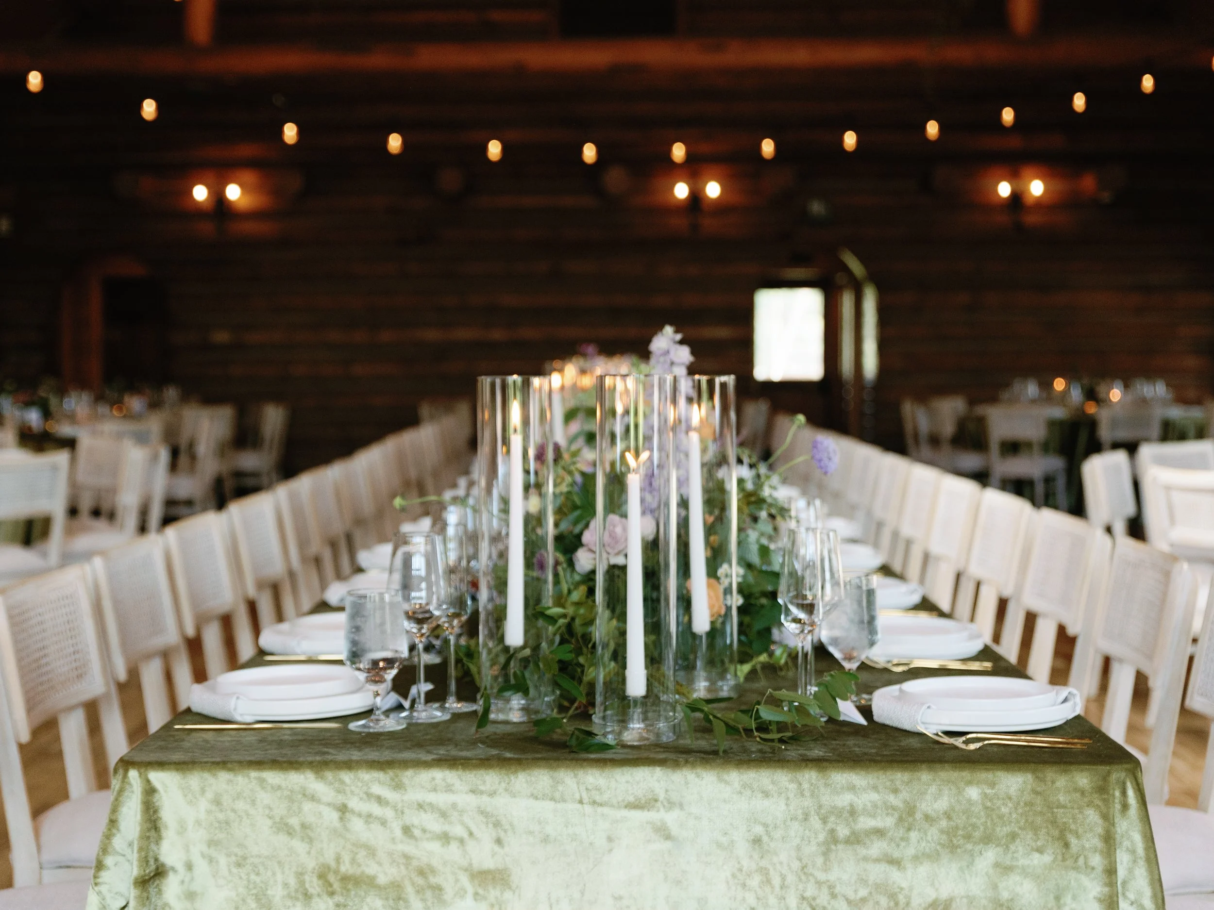 Wedding reception table set with floral centerpiece, tall candles, and elegant tableware inside a rustic wooden barn with string lights.