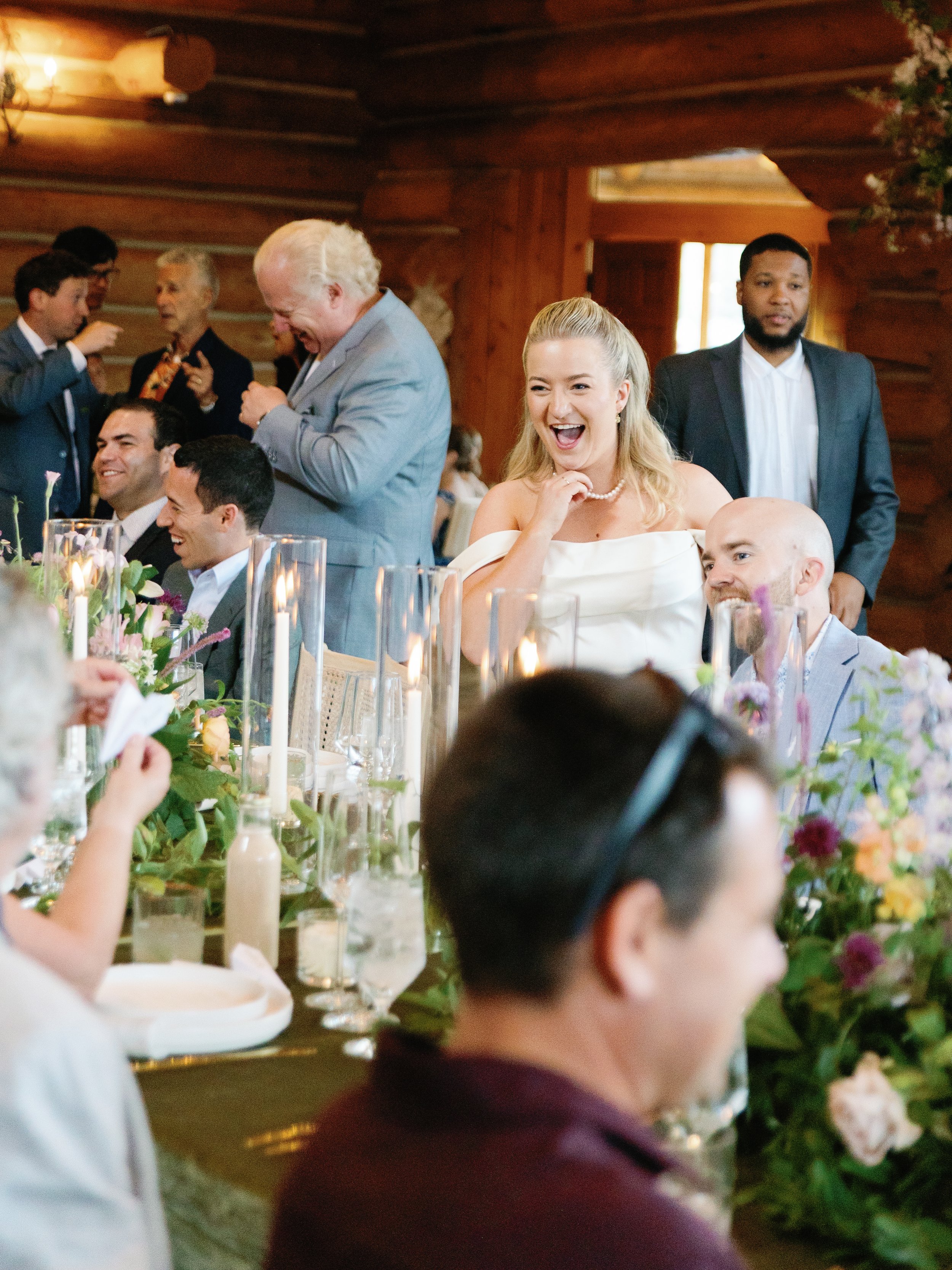 Group of people at a wedding reception, sitting at a table decorated with flowers and candles, with a rustic wood interior. Some are laughing and talking, with a woman in a white off-shoulder dress sharing a joyful moment.